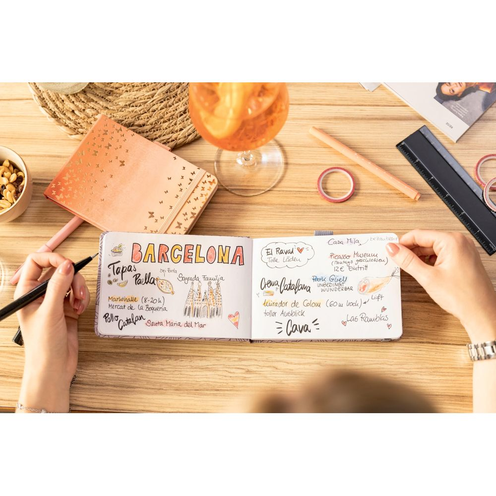 Una mano scrive su un taccuino con appunti su Barcellona. Una bevanda e snack sono nelle vicinanze.