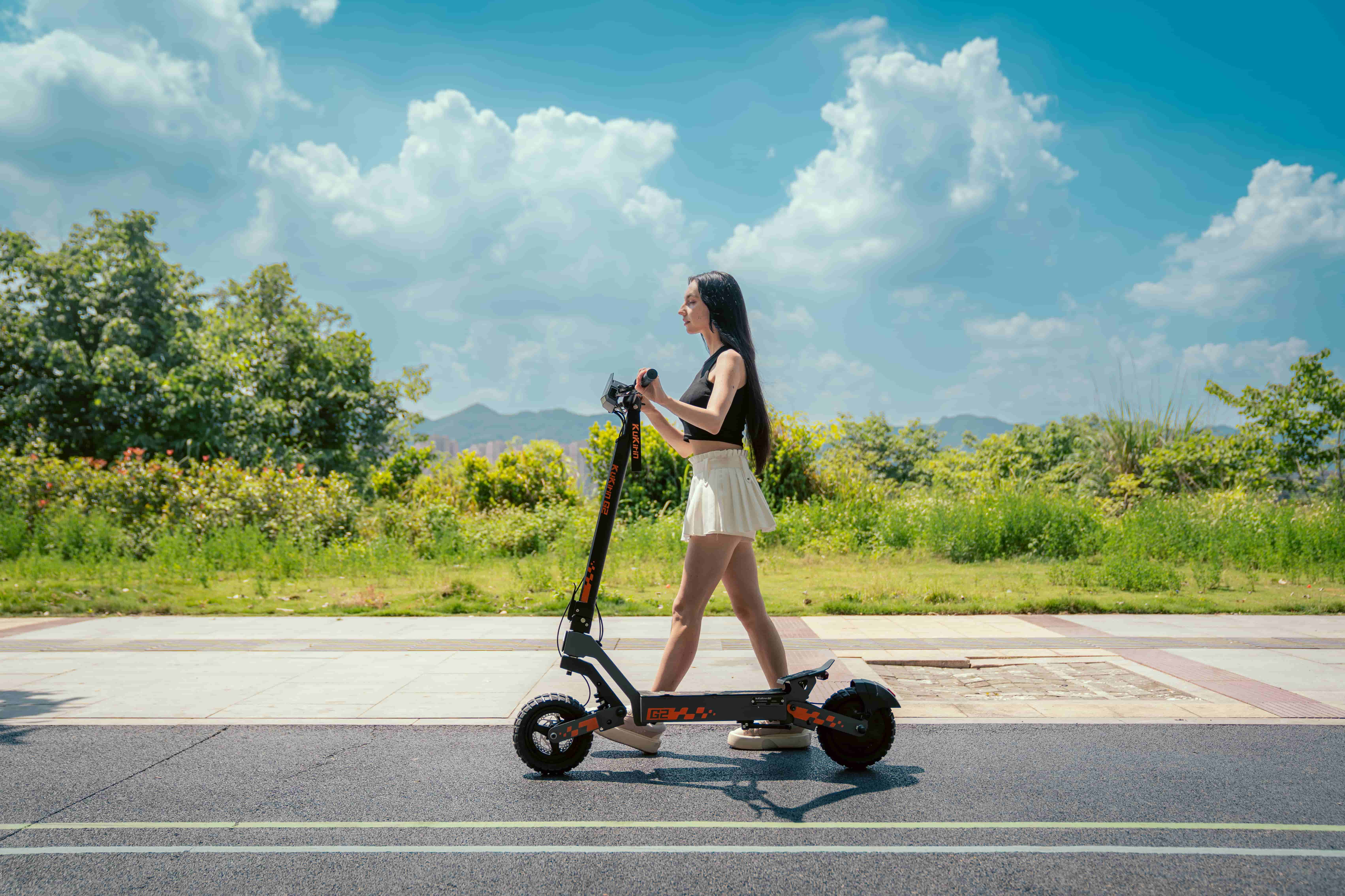 Eine Frau fährt einen schwarzen Elektroroller auf einer Straße mit grüner Vegetation und blauen Himmel.