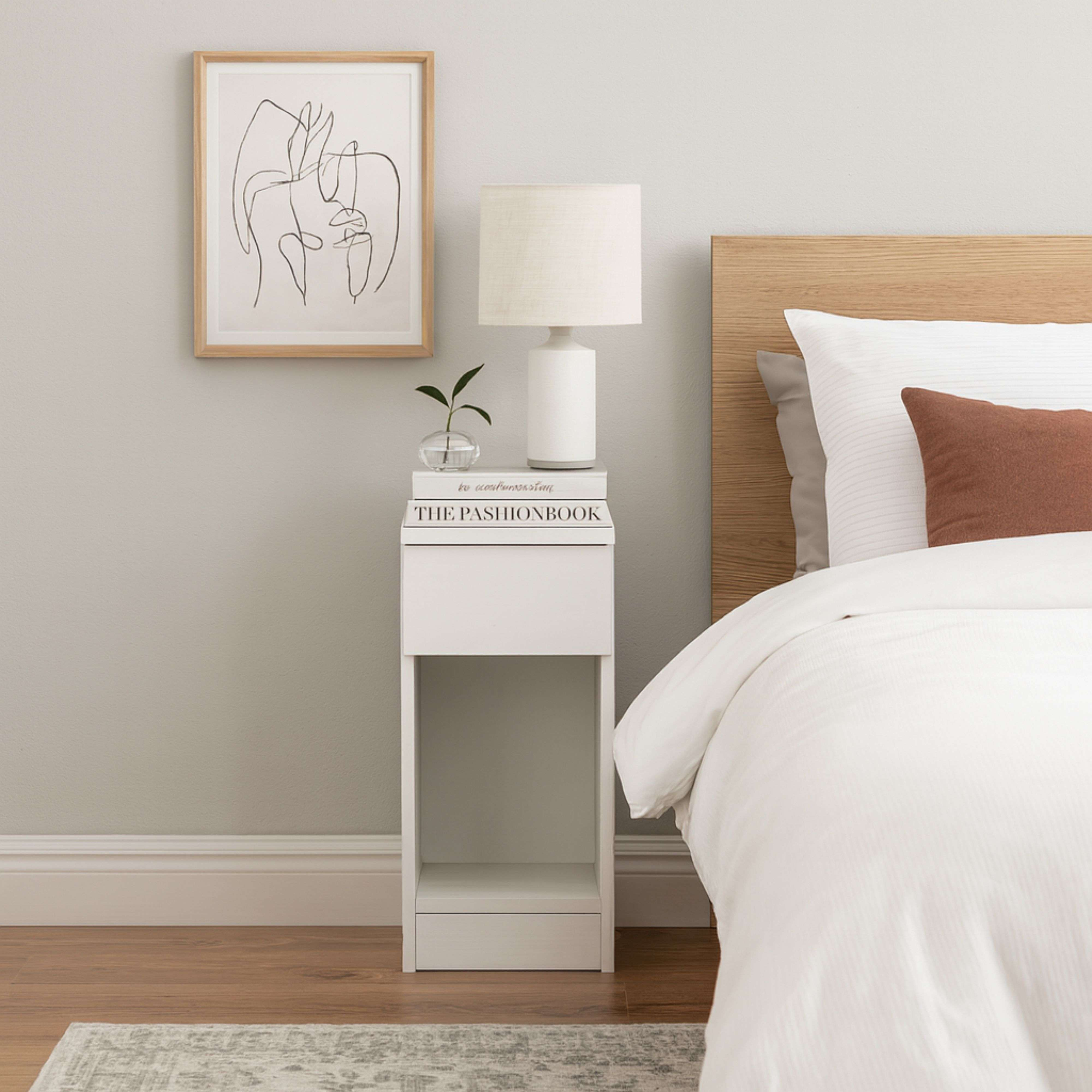 Una mesita de noche blanca con lámpara, libros y planta junto a una cama con ropa de cama blanca.