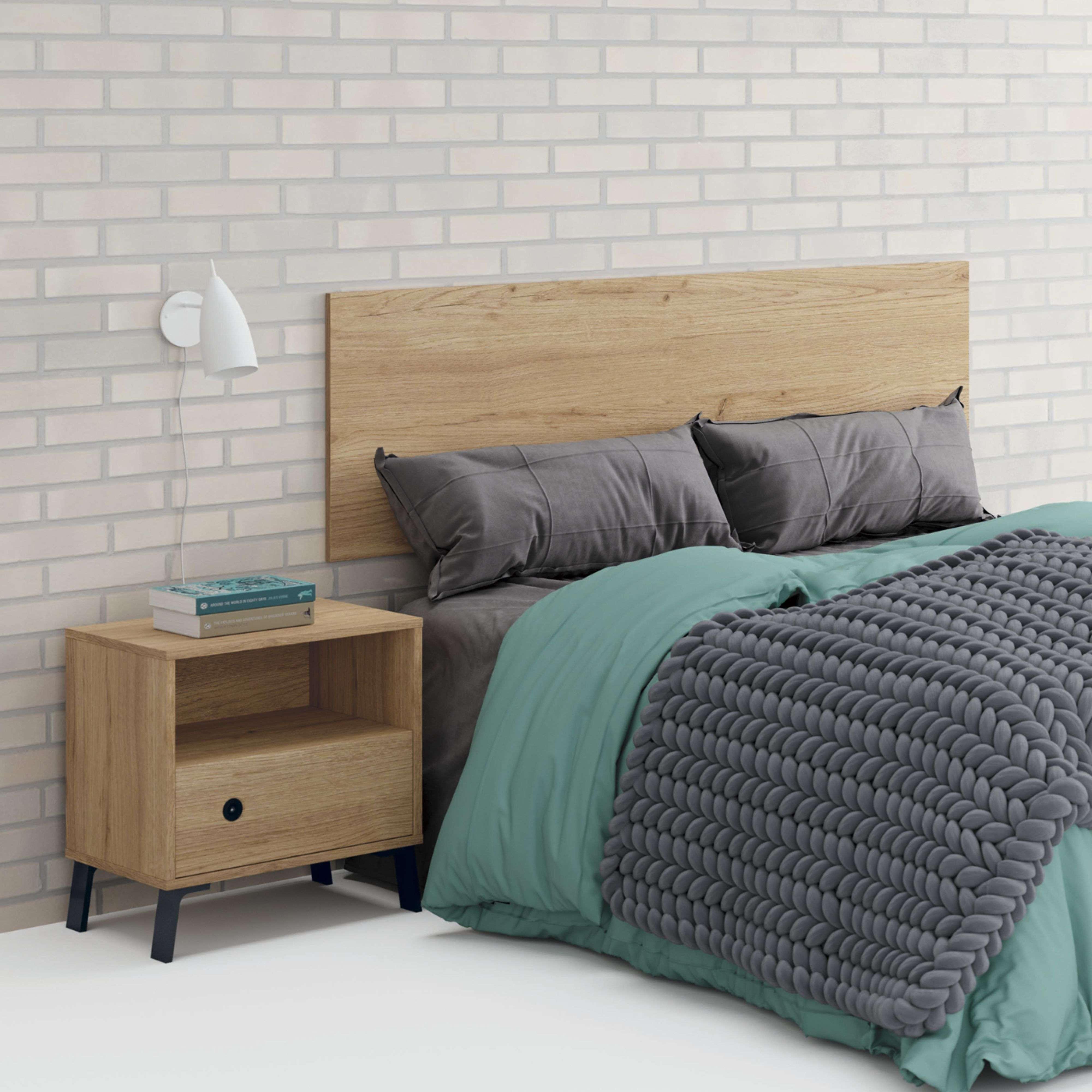 Mesita de noche de madera junto a una cama con almohadas grises, ropa de cama verde azulado y manta tejida.