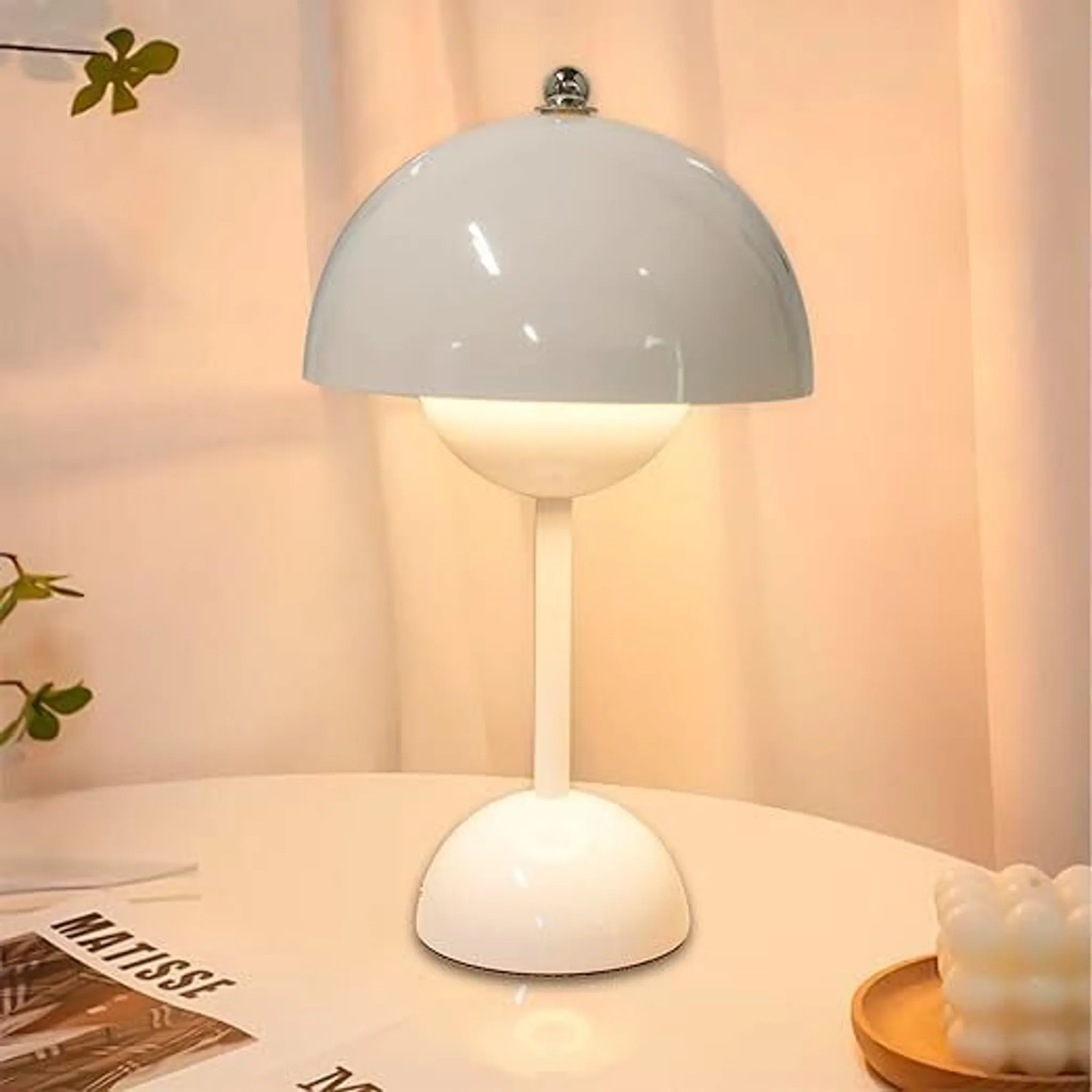 Weiße Pilzlampe mit silbernem Knopf auf einem Tisch mit Büchern und Kerze.