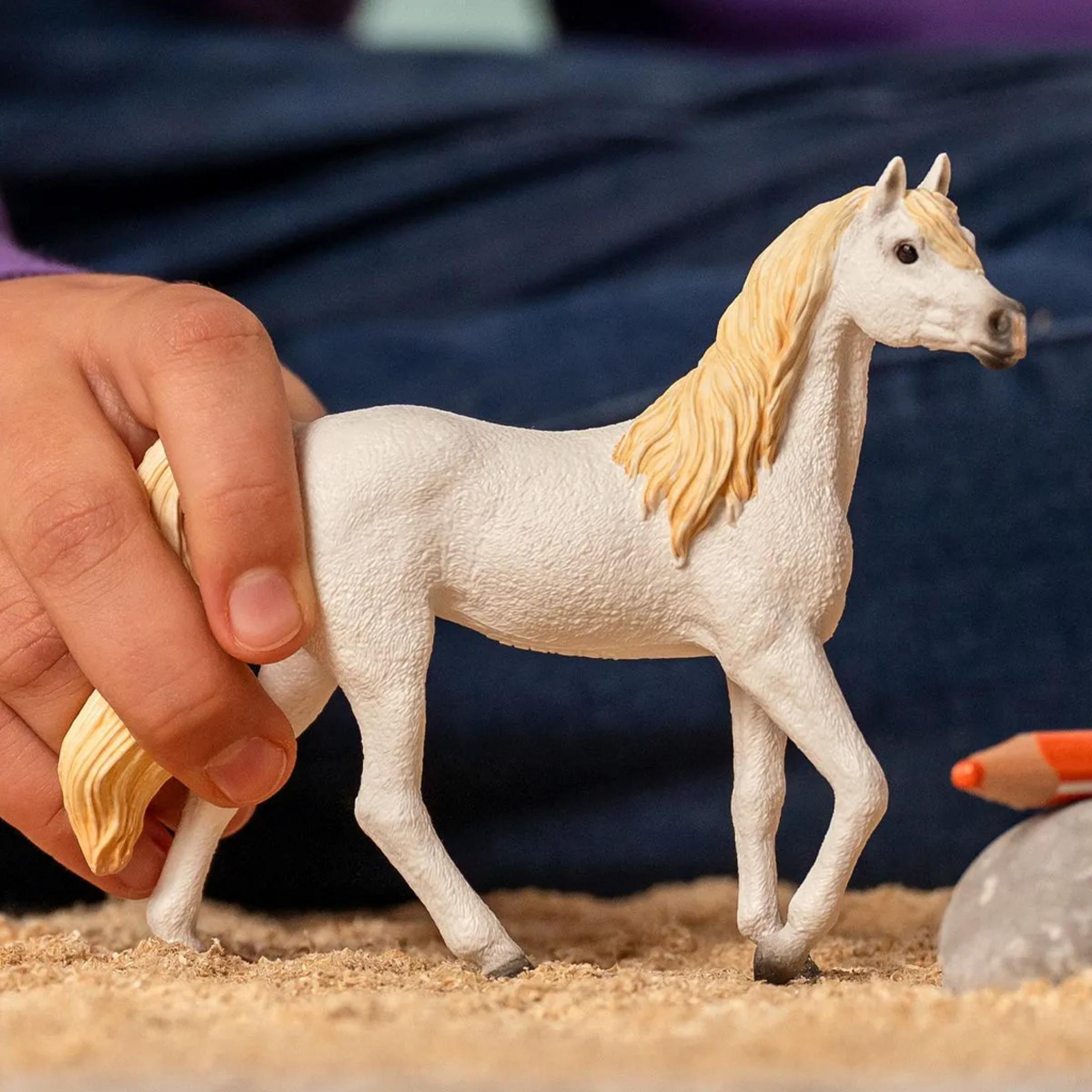 Un cavallo giocattolo bianco con criniera bionda nella sabbia, tenuto da una mano.