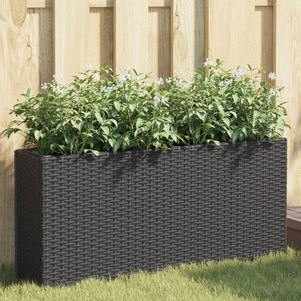 Una fioriera in rattan grigio scuro piena di piante verdi con piccoli fiori bianchi.