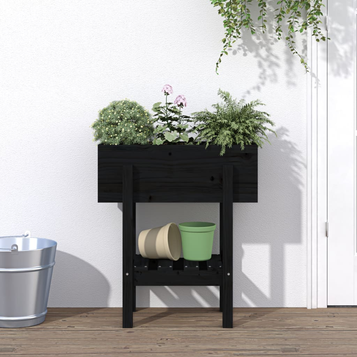 Ein schwarzer Blumenkasten auf Beinen mit Pflanzen und zwei Töpfen. Ein Eimer steht links.
