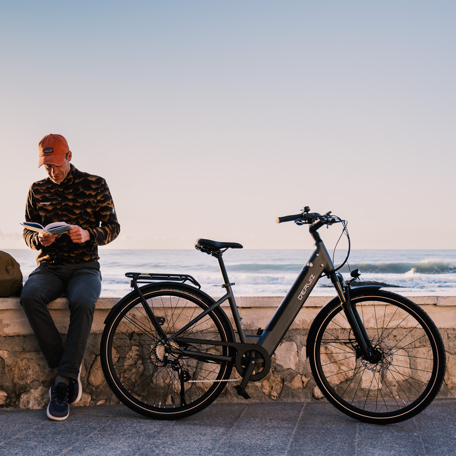 Mann mit orangefarbener Kappe liest Buch am Fahrrad, Meer und Mauer im Hintergrund.