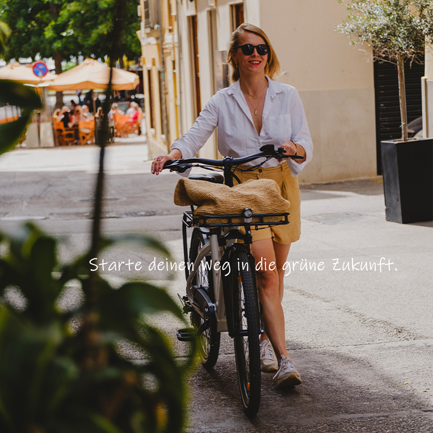 Eine Frau mit Fahrrad steht auf einer Straße mit Cafés im Hintergrund.