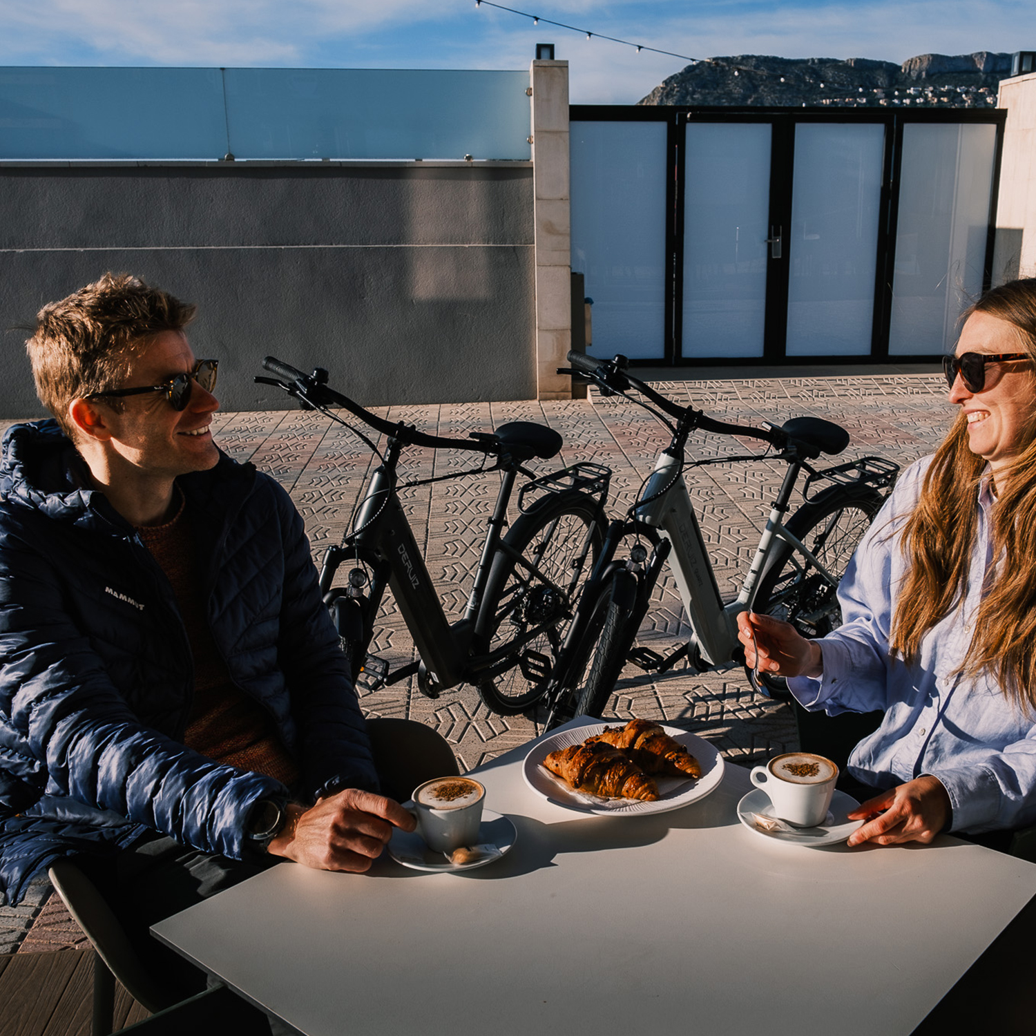 Mann und Frau sitzen an einem Tisch mit Kaffee und Croissants, Fahrräder im Hintergrund.