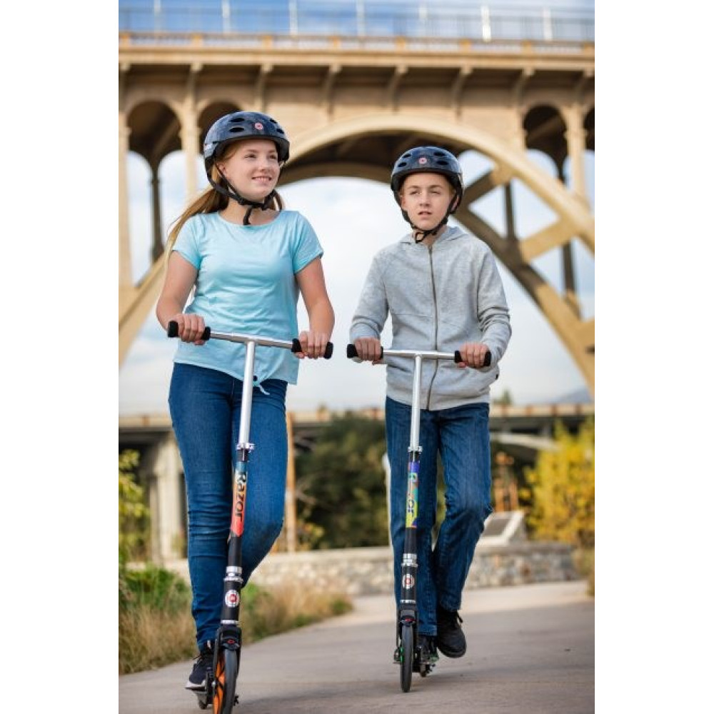 Two children wearing helmets ride scooters under a bridge.