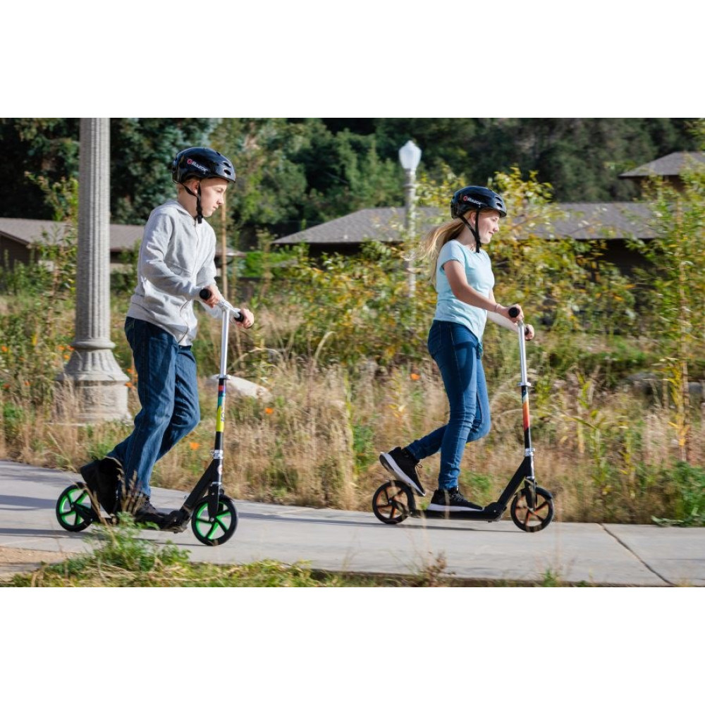 Two children ride scooters on a paved path in a park-like setting.