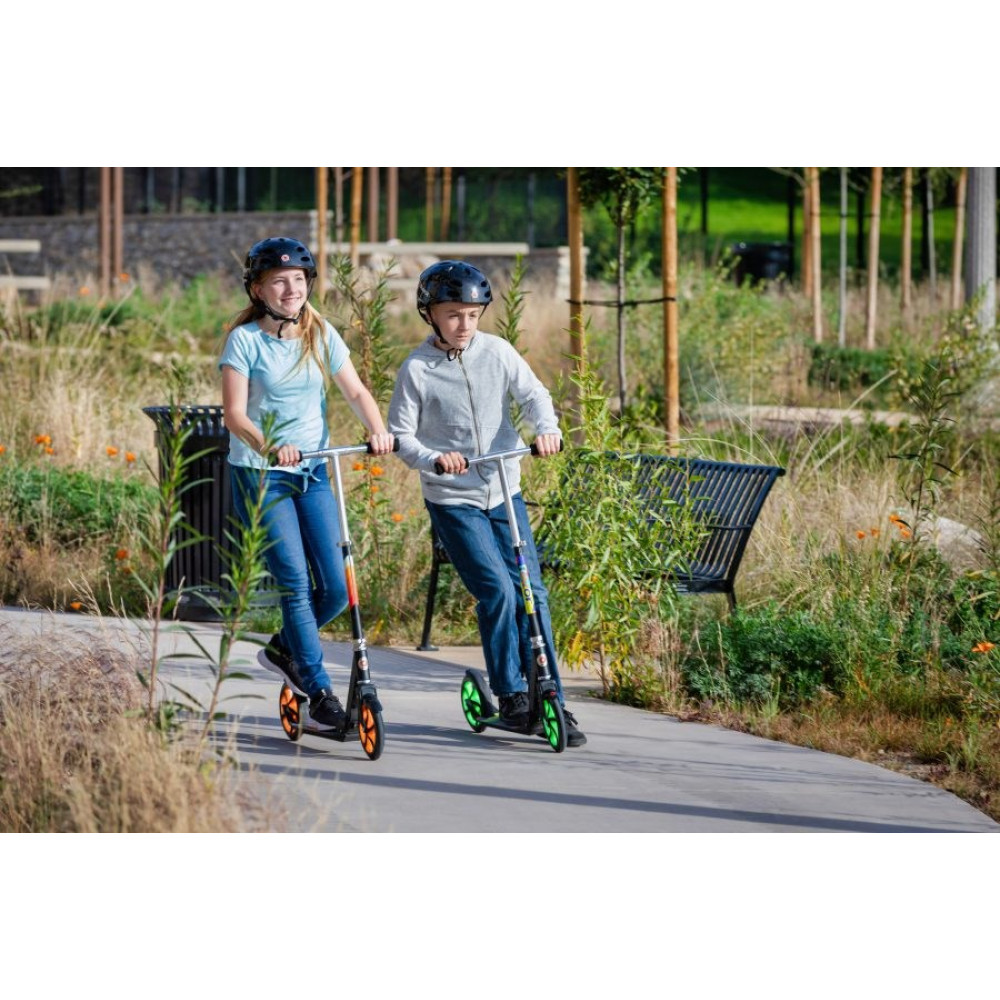 Two kids with helmets ride scooters on a path in a park with lush greenery.