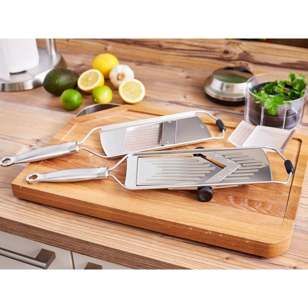 Due mandoline su un tagliere di legno con frutta e aglio sullo sfondo.