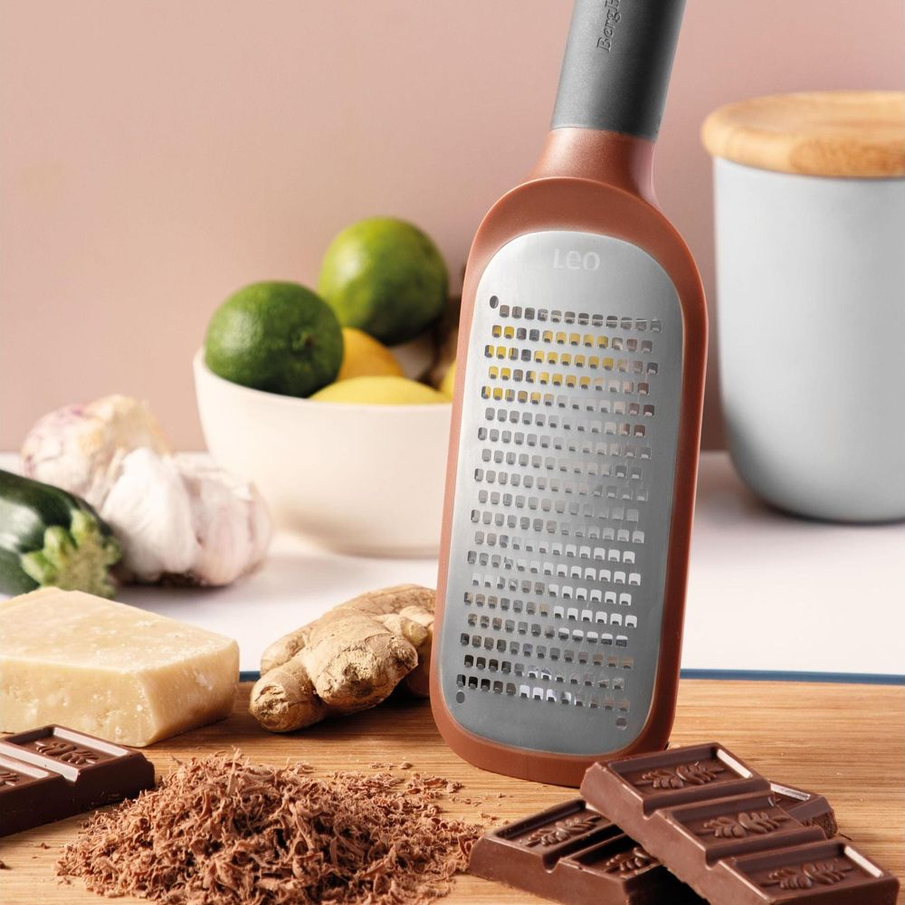 Una grattugia con manico marrone giace su un tagliere di legno con cioccolato, zenzero, formaggio e agrumi.