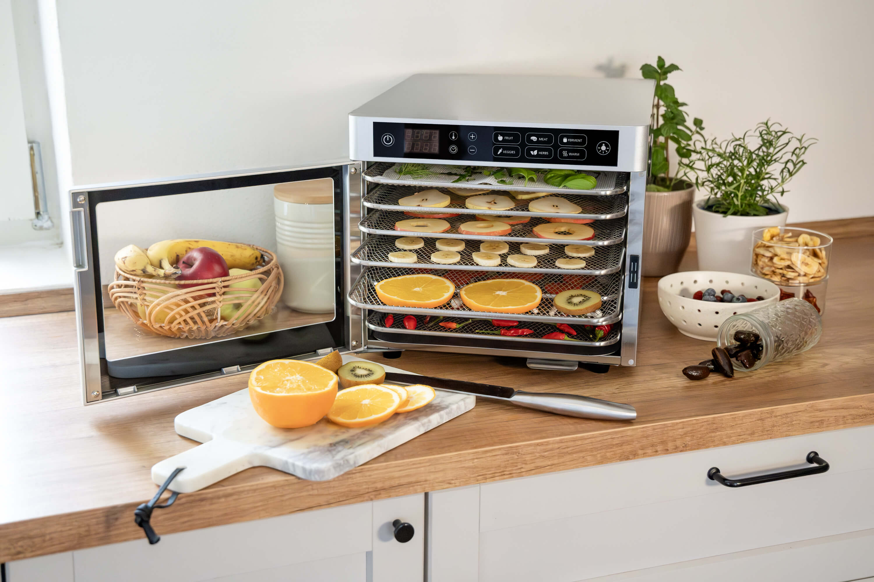Ein Dörrgerät mit Obst, Kräutern und Chilis auf den Rosten. Daneben Obst und Beeren in Schalen.