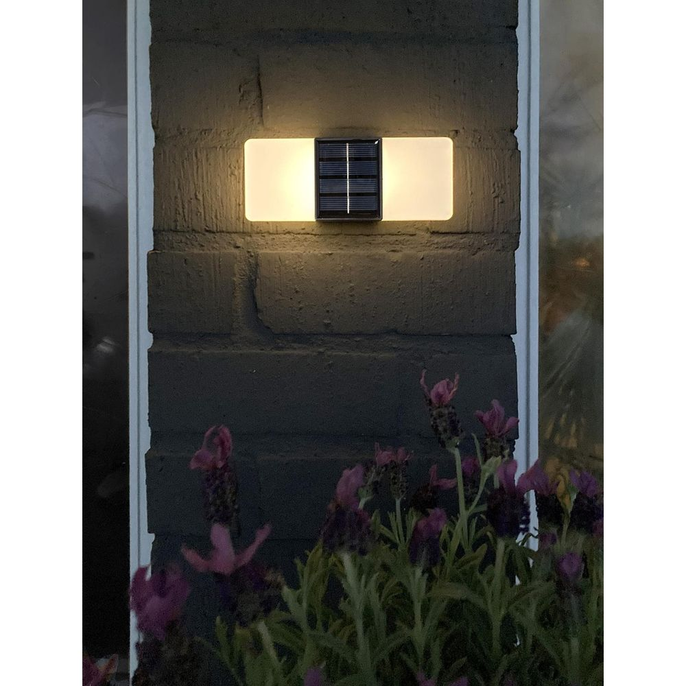 Solarleuchte an dunkler Ziegelwand, beleuchtet weiße Rechtecke, mit lila Blumen im Vordergrund.