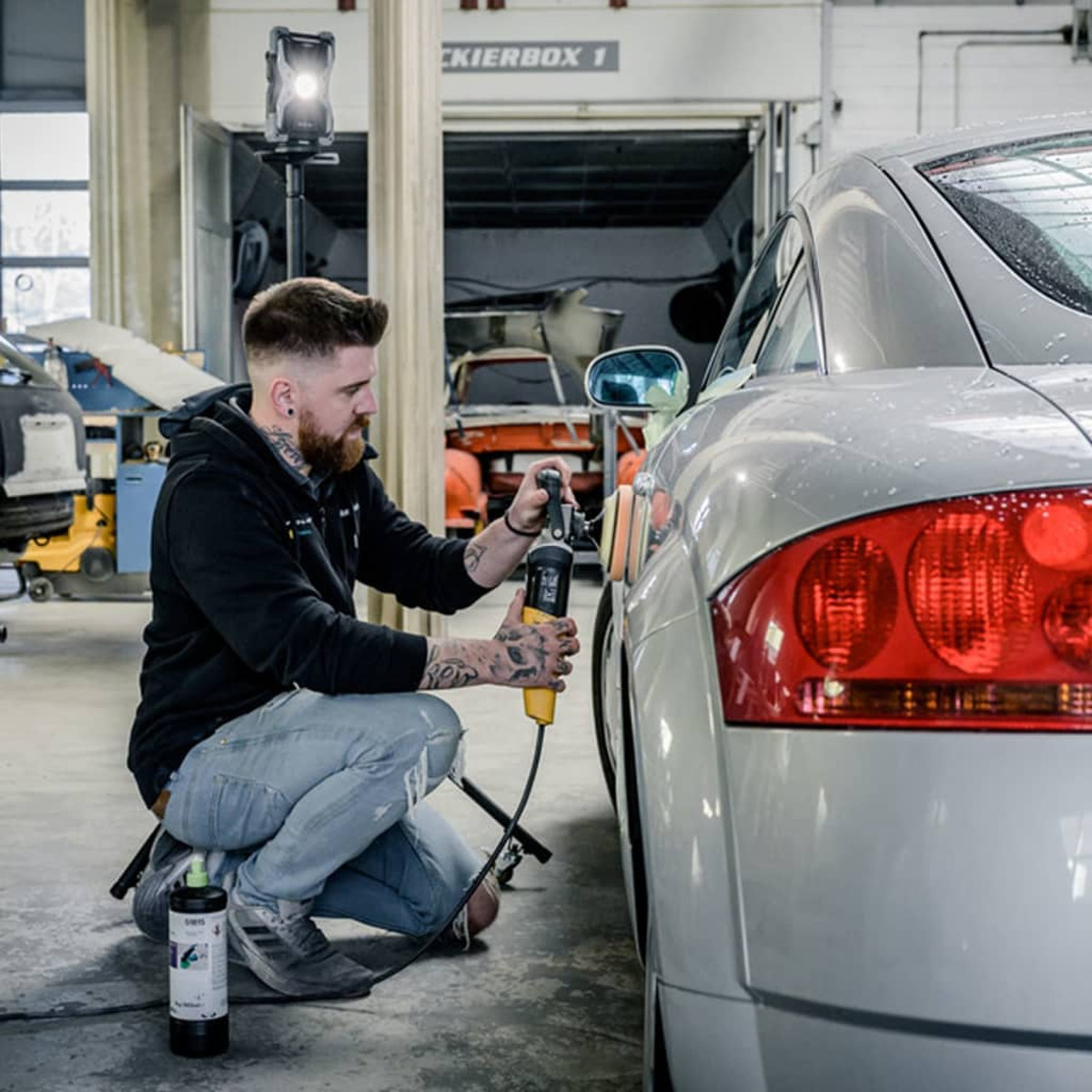 Mann im schwarzen Hoodie poliert silbernes Auto mit gelbem Polierer in Garage.