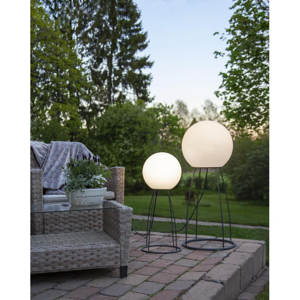 Zwei leuchtende Kugelleuchten auf Ständern, eine Gartenlounge mit Decke und Pflanzen.