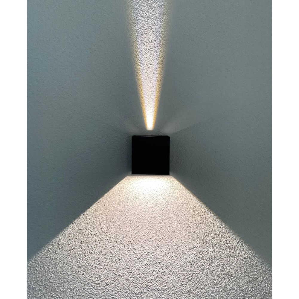 Applique murale cubique noire projetant des faisceaux lumineux vers le haut et vers le bas sur un mur texturé.