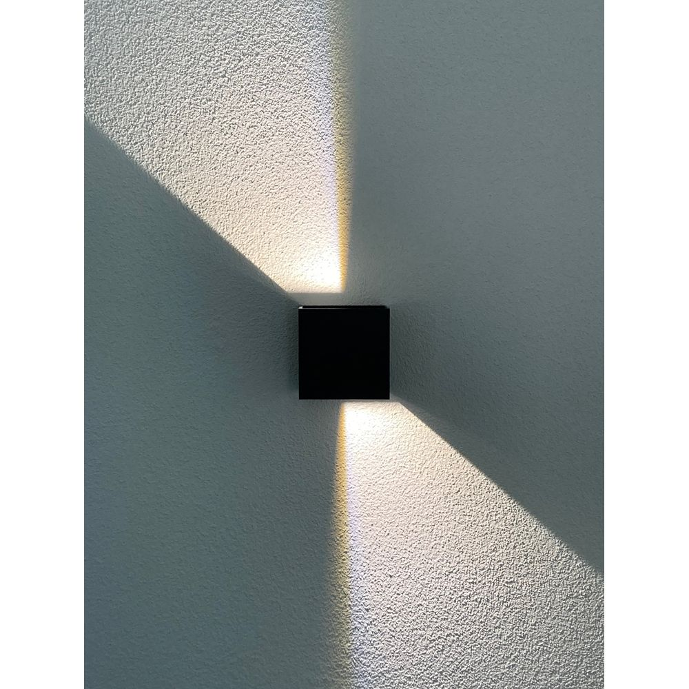 Un applique mural cubique noir éclaire vers le haut et vers le bas sur un mur blanc texturé.