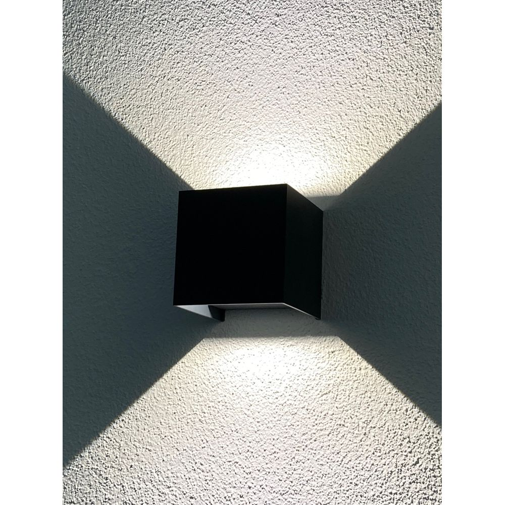 Un applique mural extérieur noir, en forme de cube, projette des faisceaux de lumière symétriques.