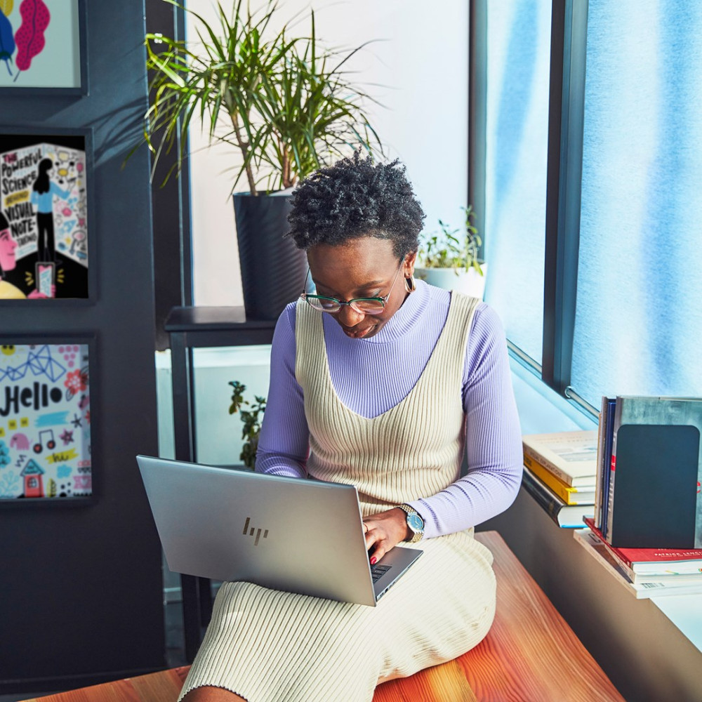 Kobieta w okularach pracuje na laptopie, siedząc na drewnianej ławce z roślinami i sztuką na ścianie.