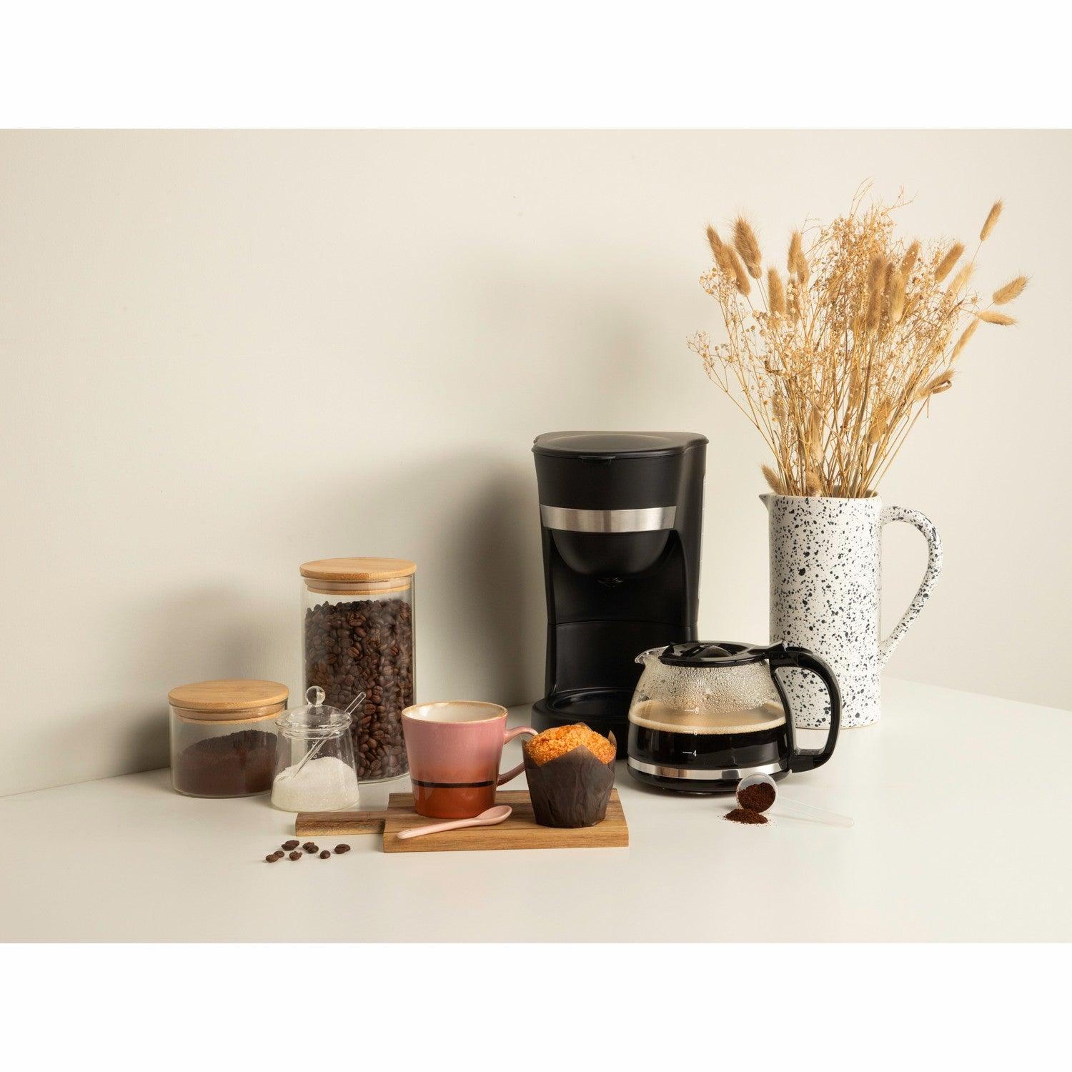 Koffiezetapparaat met kan, mok, muffin, koffiebonen en potten op een tafel.