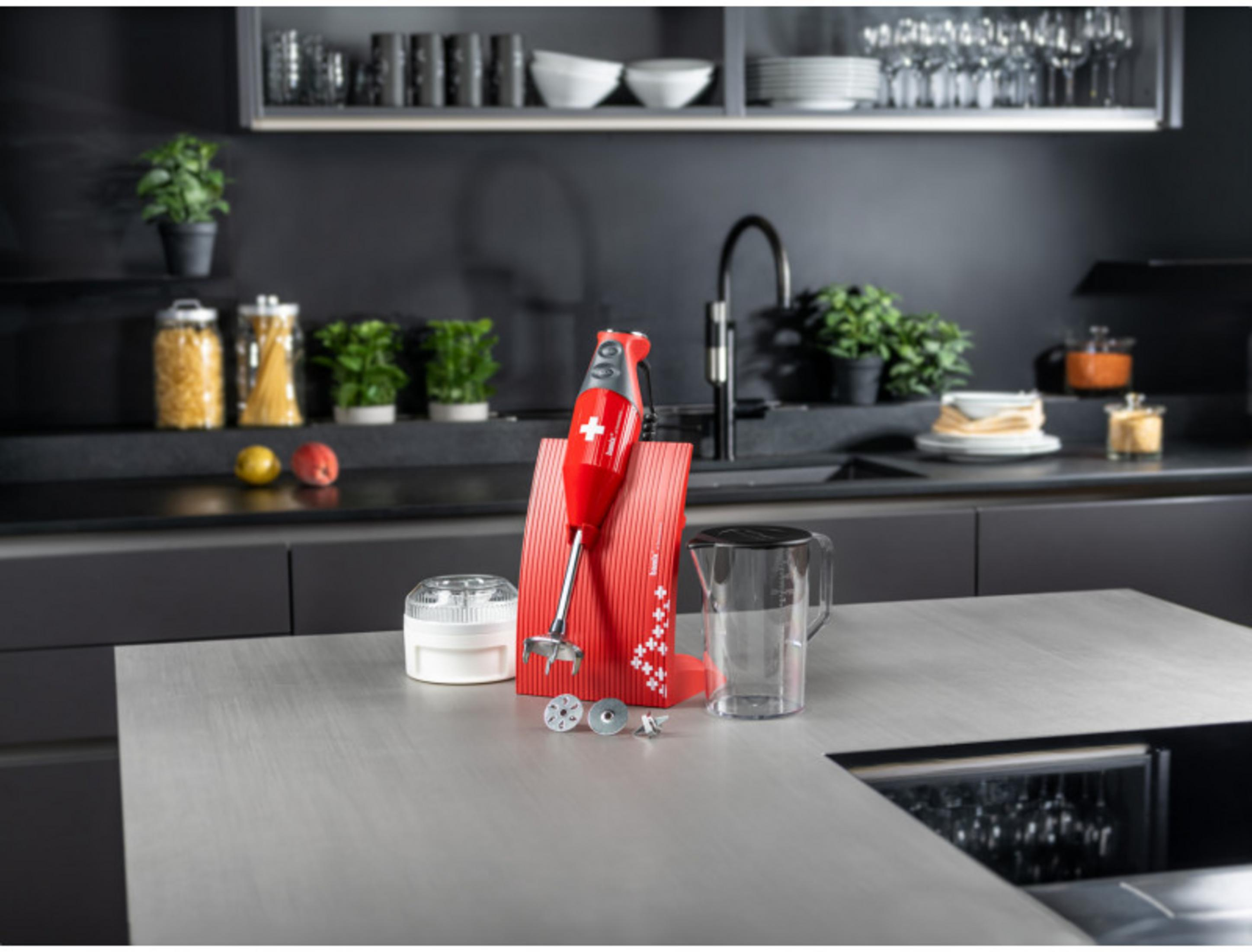 Ein roter Handmixer mit Zubehör steht auf einer Arbeitsplatte neben einem Krug.