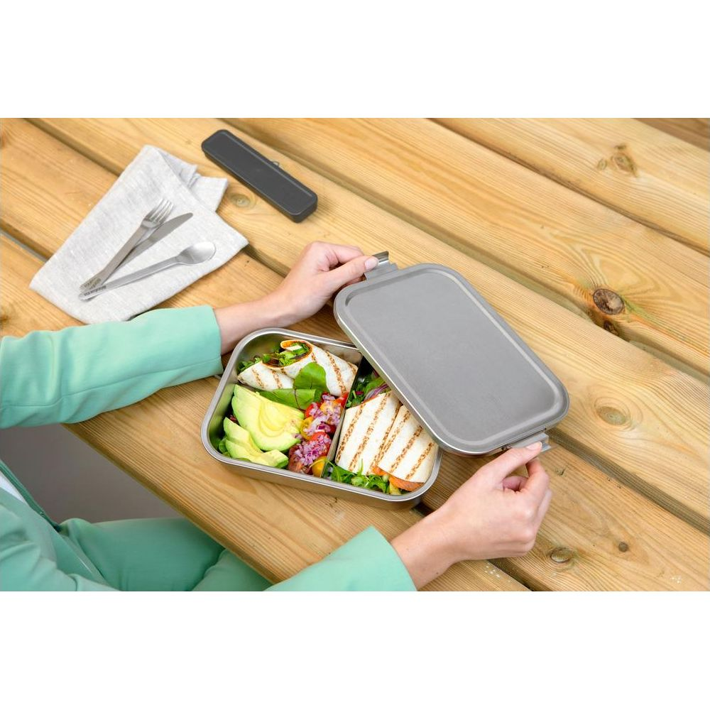 Una persona apre un portapranzo in metallo con wrap e avocado su tavolo di legno.