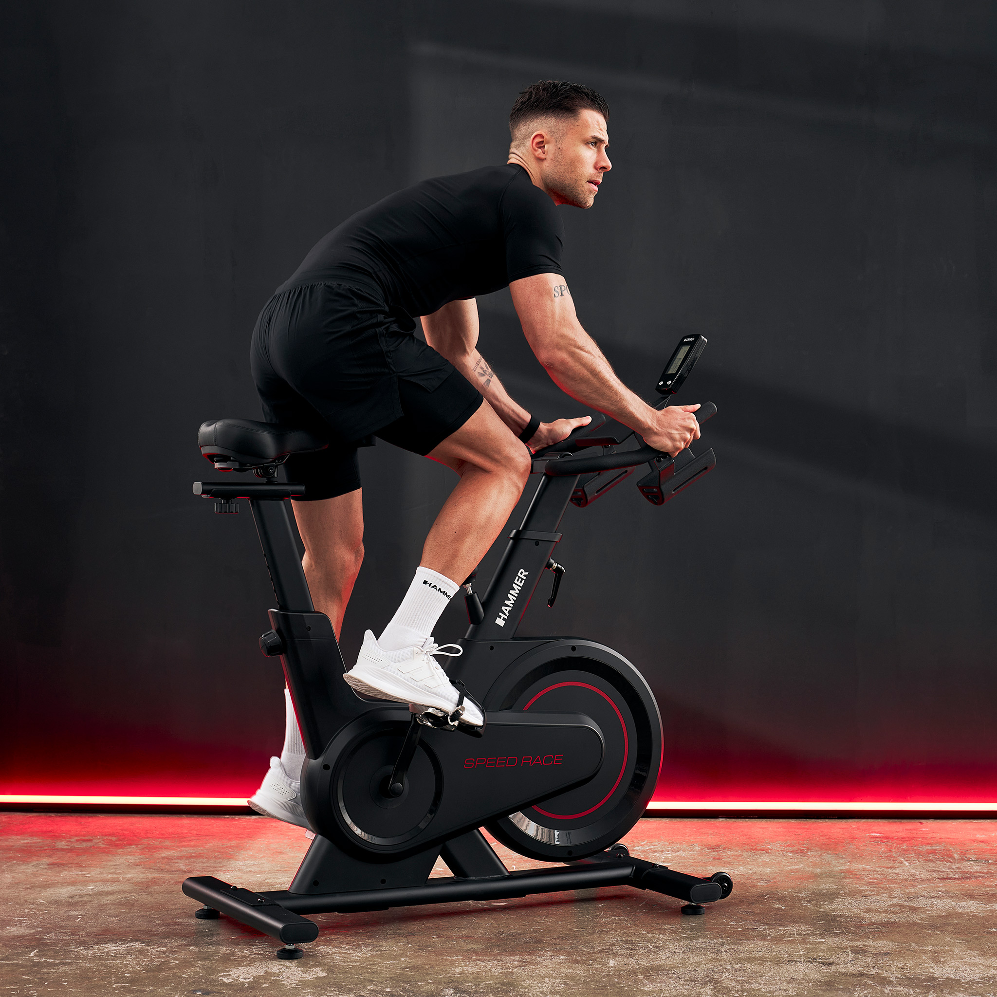 Mężczyzna na czarnym rowerze stacjonarnym z czerwonymi akcentami, ubrany na sportowo.