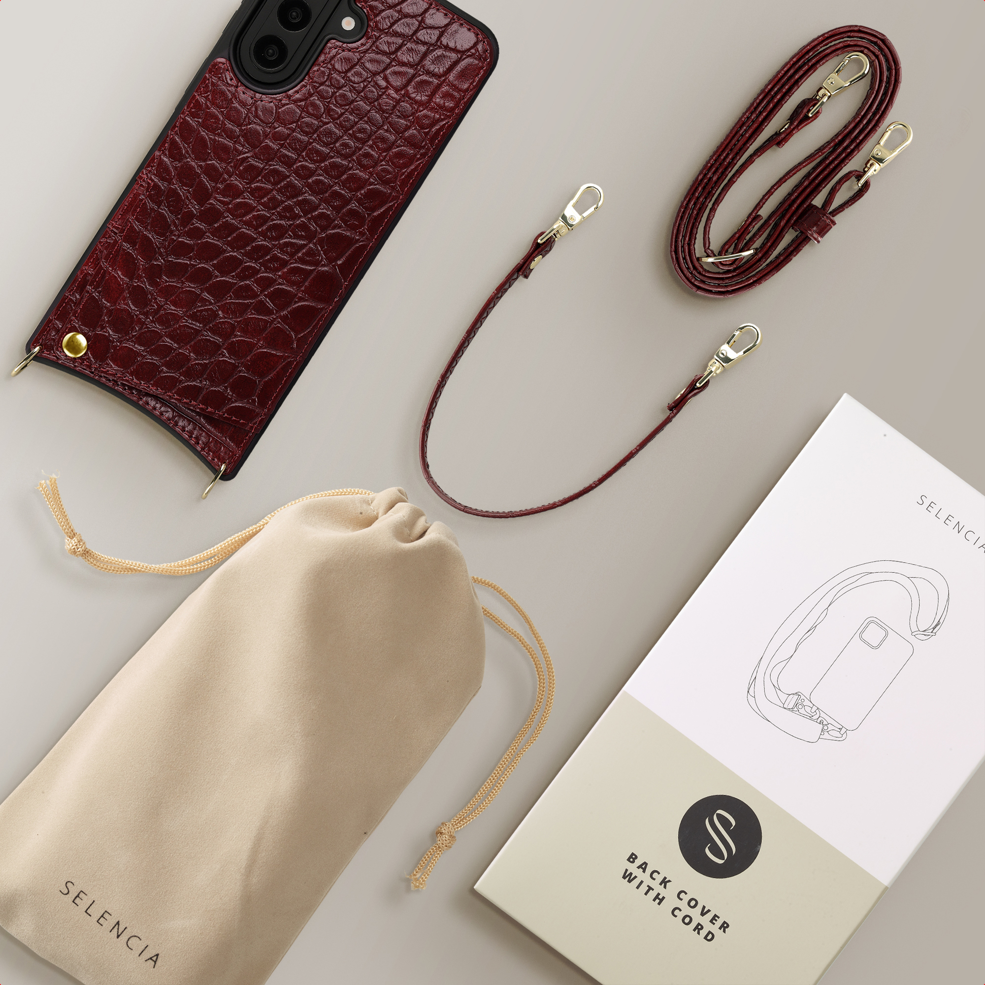 Una custodia per telefono in rilievo di coccodrillo rosso scuro, cordino, custodia e scatola sono disposti su una superficie grigio chiaro.