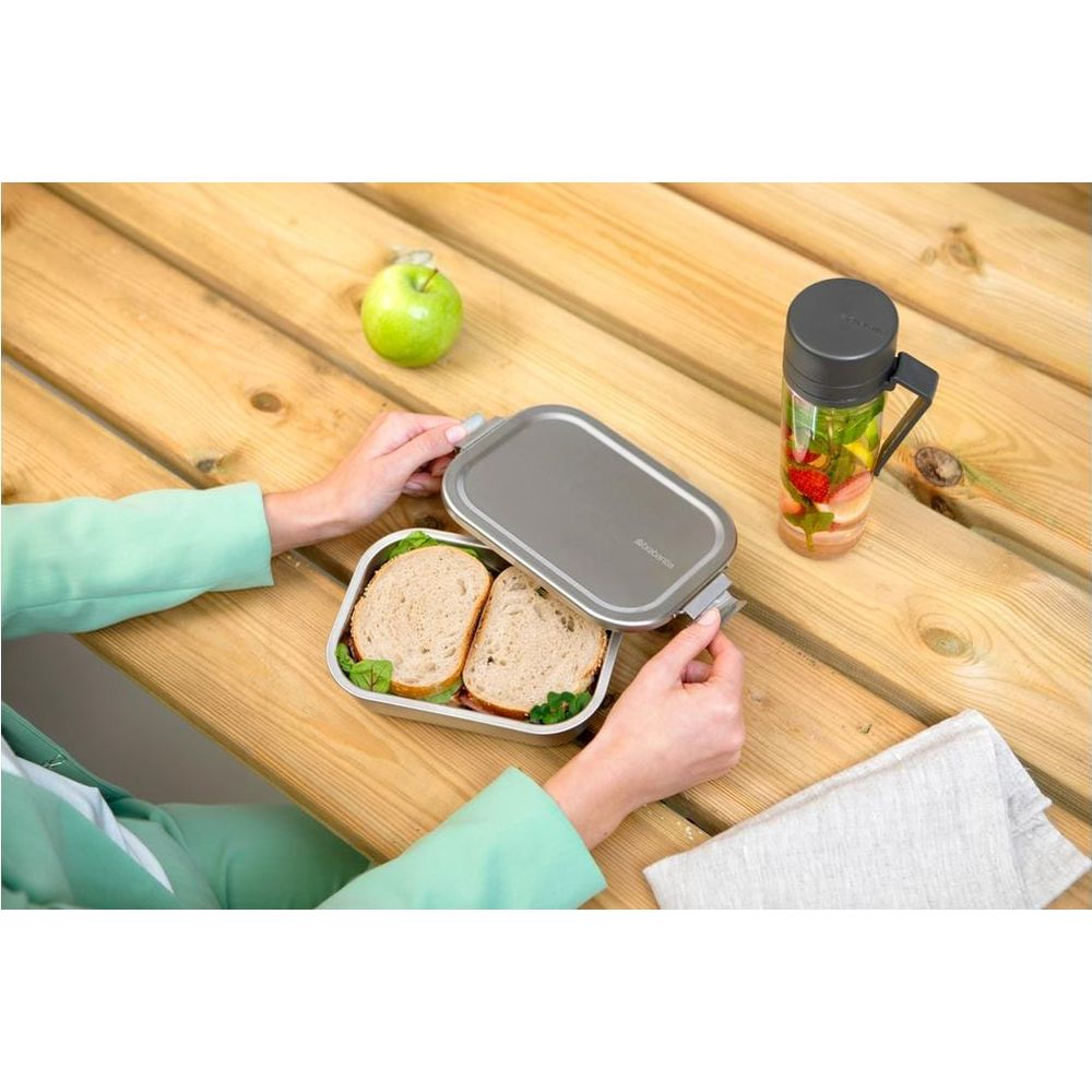 Una persona apre un portapranzo con due panini su un tavolo di legno.