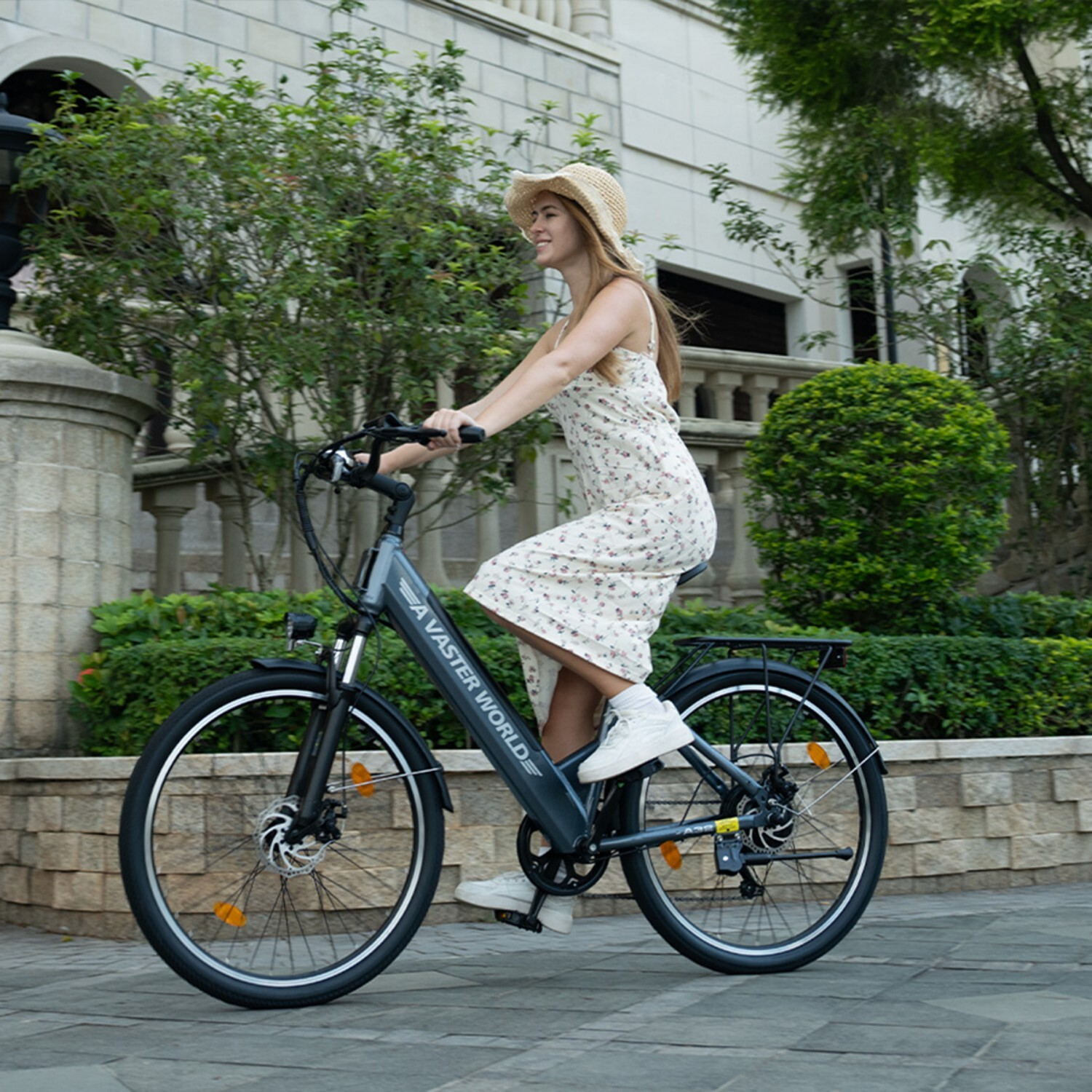 Kobieta w kapeluszu słomkowym jedzie na rowerze elektrycznym w kwiecistej sukience, drzewa w tle.
