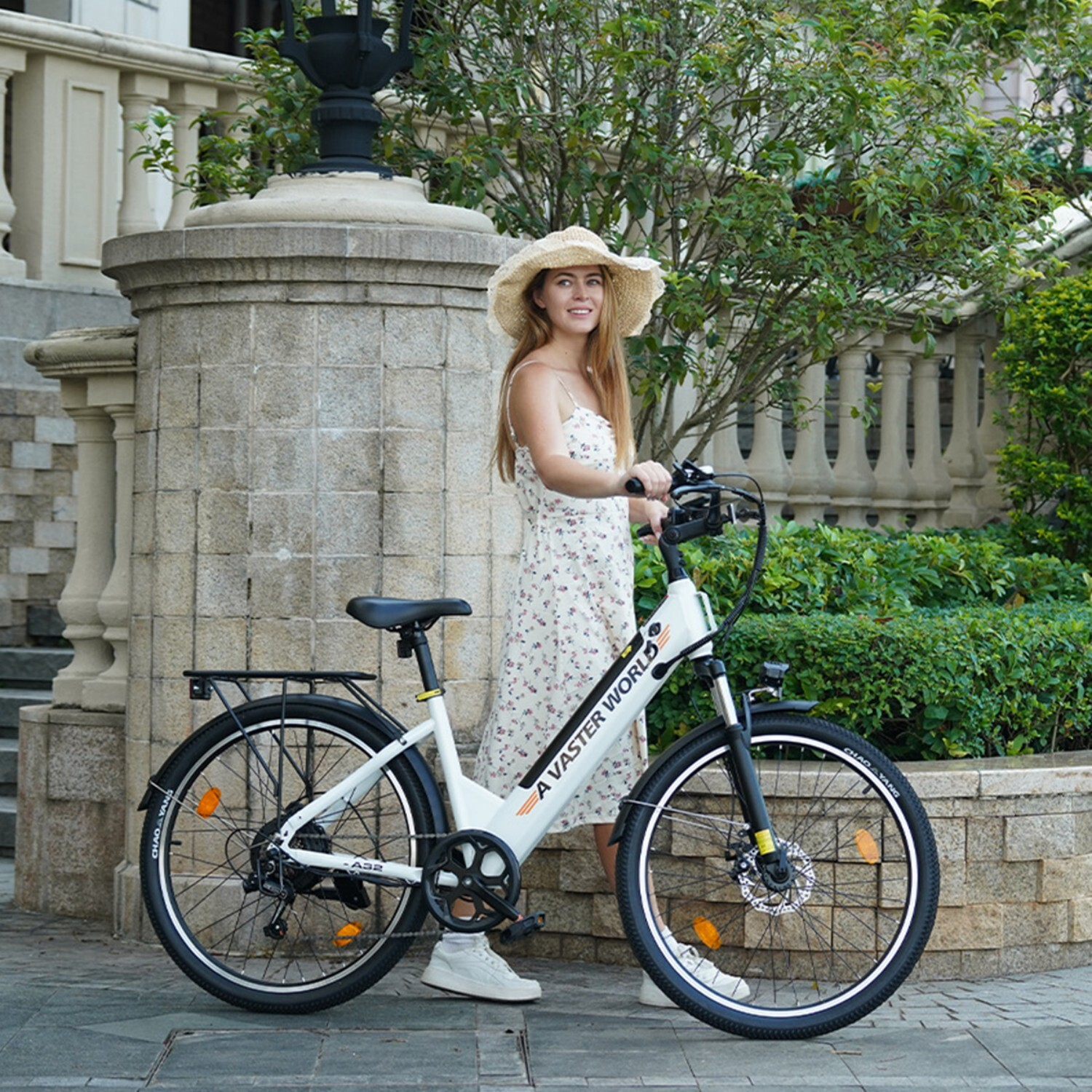 Kobieta w słomkowym kapeluszu i sukience stoi obok białego roweru przed kamiennym budynkiem.