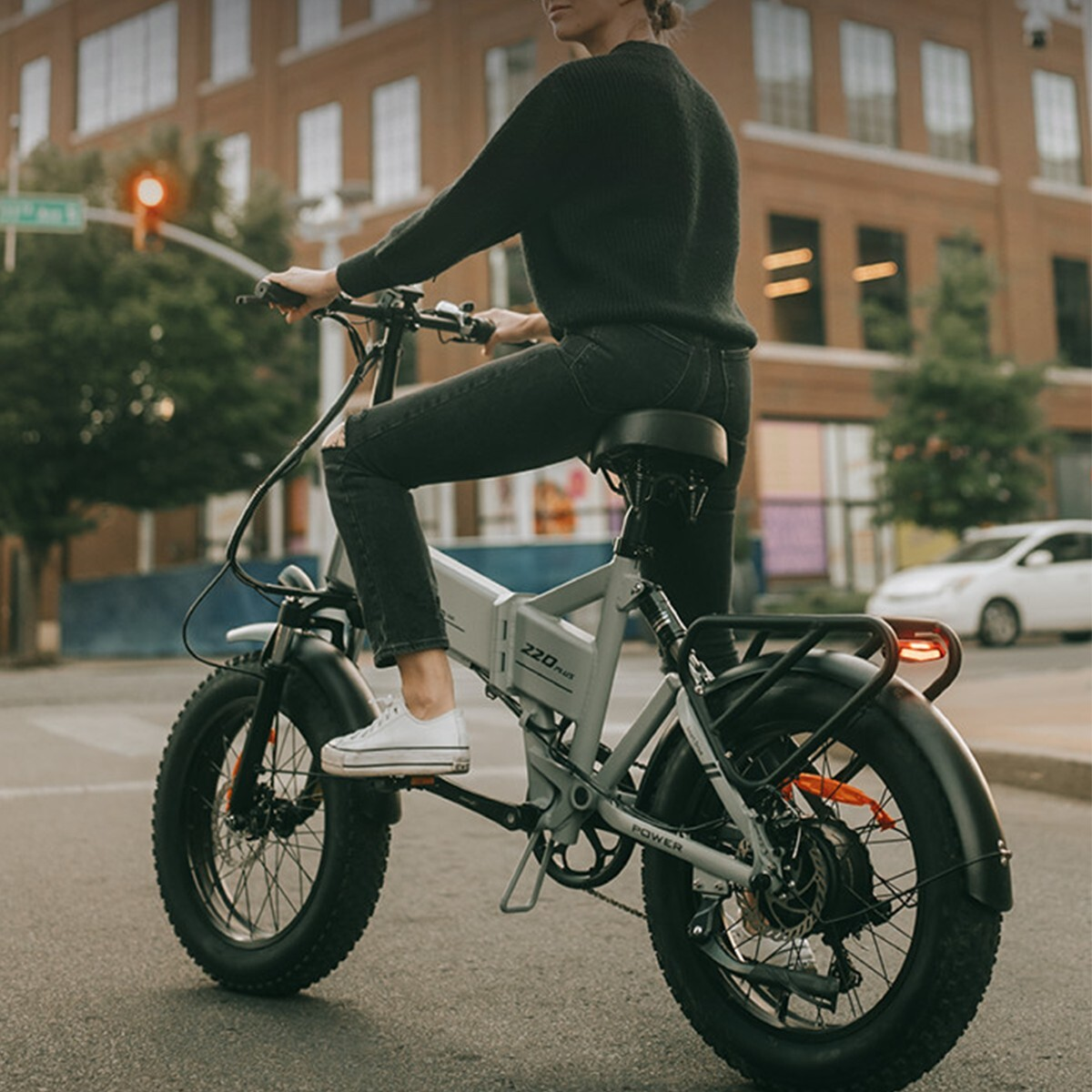 Osoba w czarnym swetrze i dżinsach jedzie szarym rowerem elektrycznym ulicą.