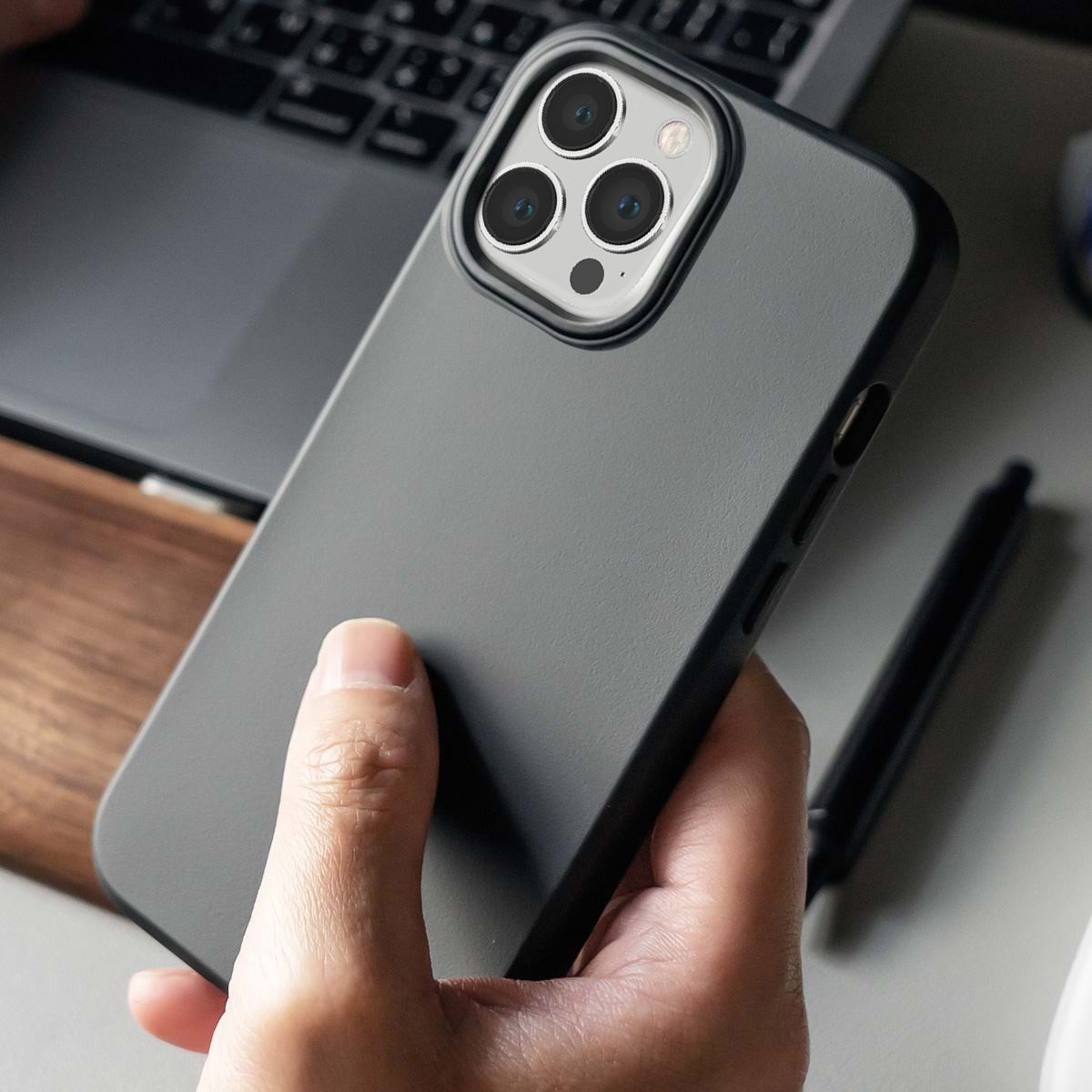 Custodia grigia per telefono con tre fotocamere tenuta da una mano. Laptop sullo sfondo.