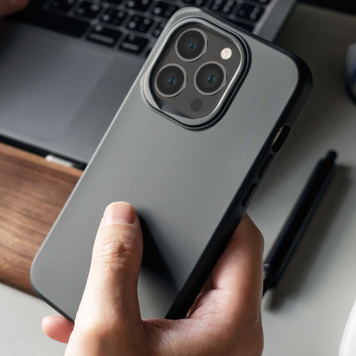 Una mano tiene un iPhone grigio scuro con tripla fotocamera, vicino a un laptop.