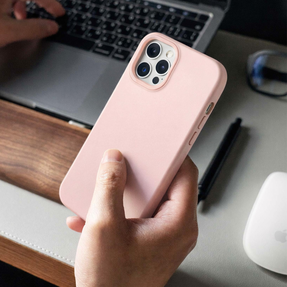 Een hand houdt een roze iPhonehoes vast bij een laptop, pen en muis.