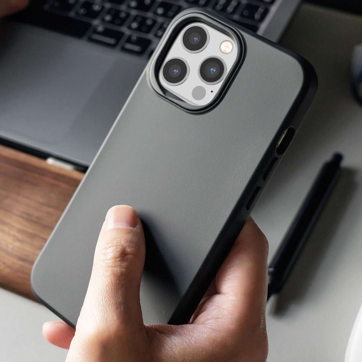 Een persoon houdt een donkergrijze telefoonhoes met drievoudige camera vast. Een laptop is erachter.