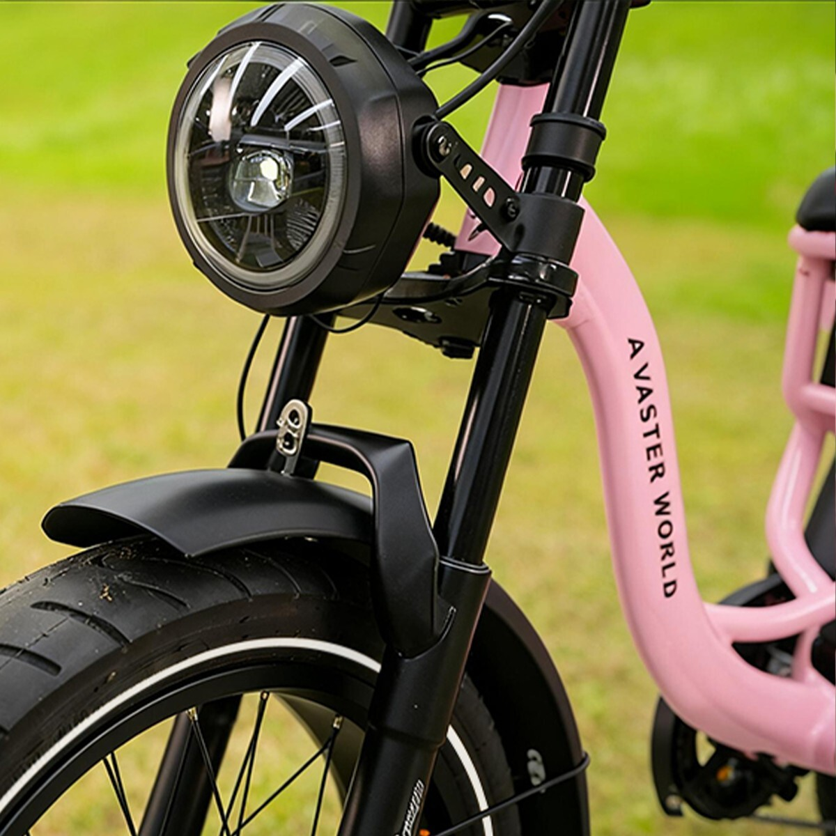 Różowy rower elektryczny z dużym okrągłym reflektorem i czarnym błotnikiem na trawie.