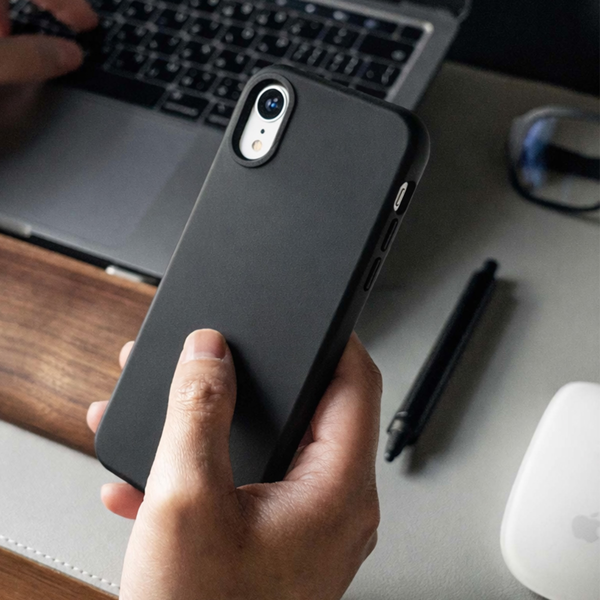 Una mano tiene uno smartphone nero davanti a un laptop, penna e mouse.