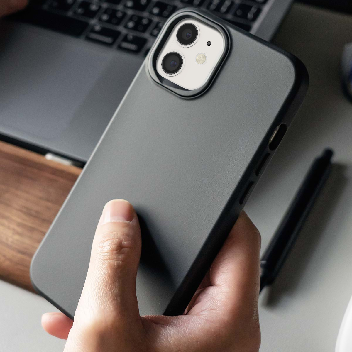 Une main tient un iPhone gris avec un double appareil photo. Un ordinateur portable est à l'arrière-plan.