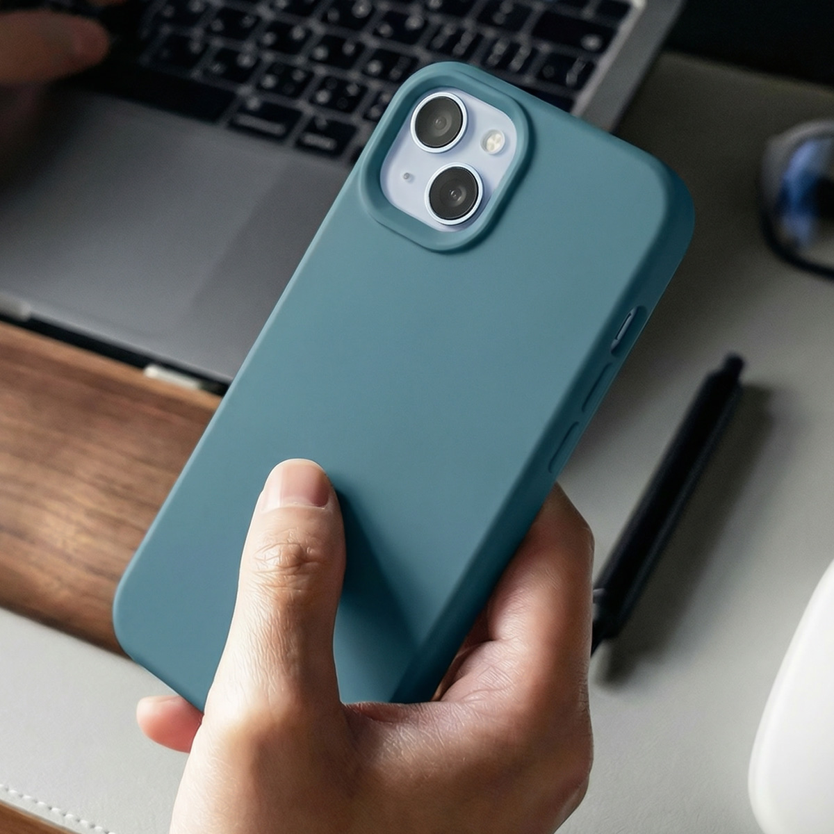 Une main tient un téléphone bleu sarcelle avec des caméras. Un ordinateur portable est en arrière-plan.