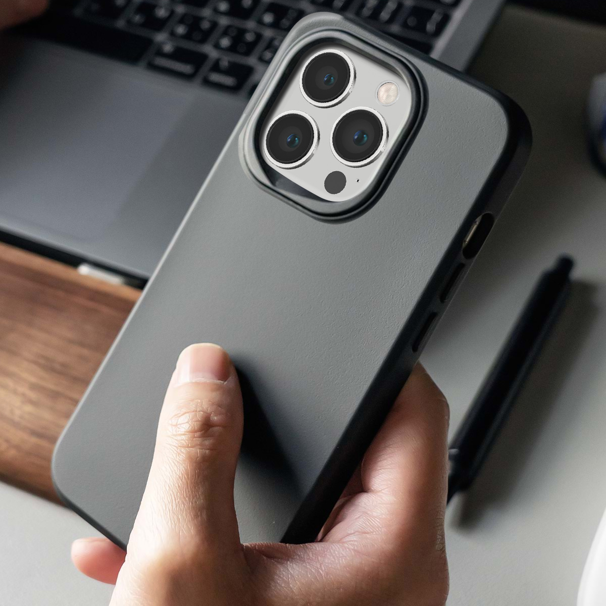 Una mano tiene una custodia grigia scura per telefono con tripla fotocamera. Un laptop sullo sfondo.