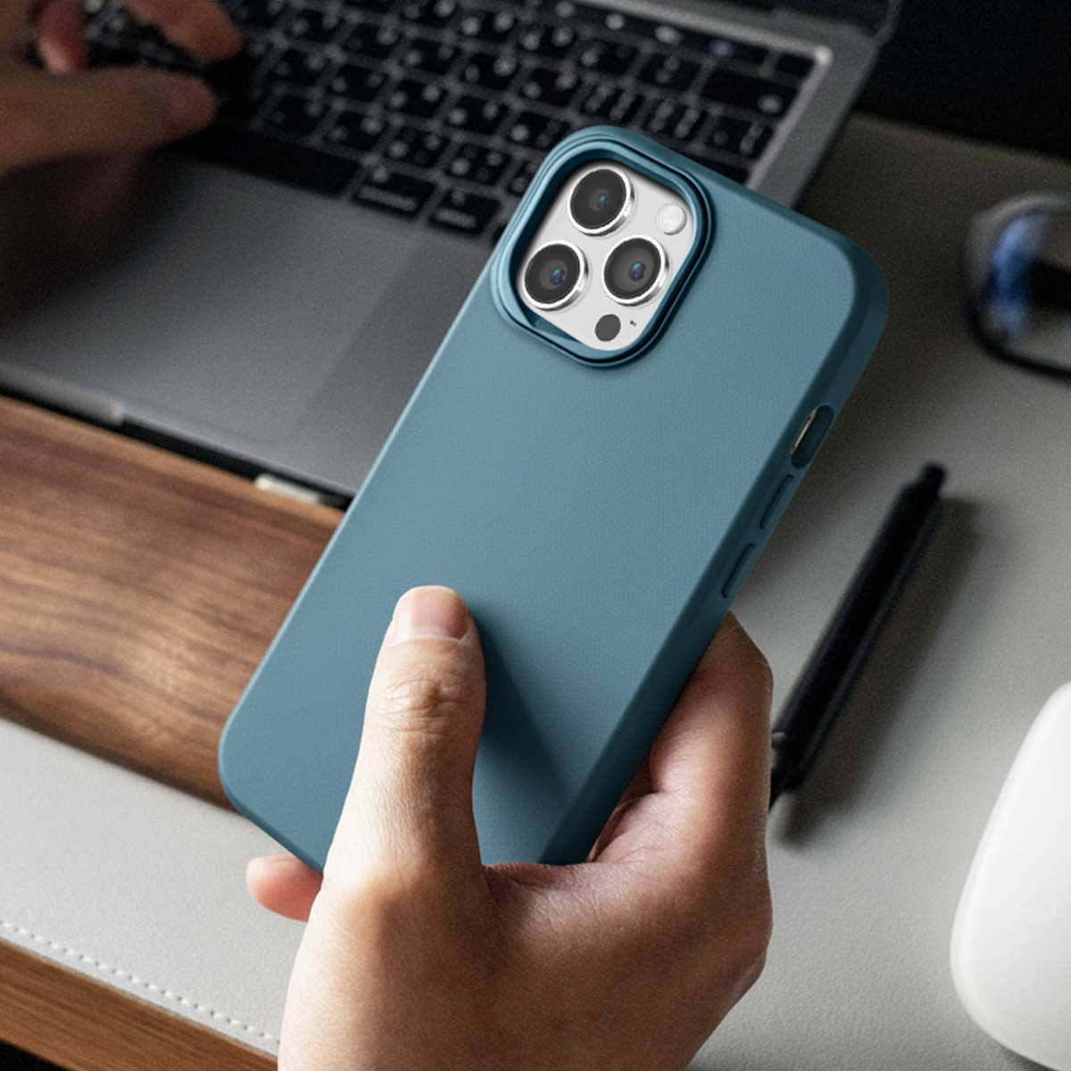 Una mano tiene un telefono blu con tre fotocamere, accanto a un laptop e una penna.