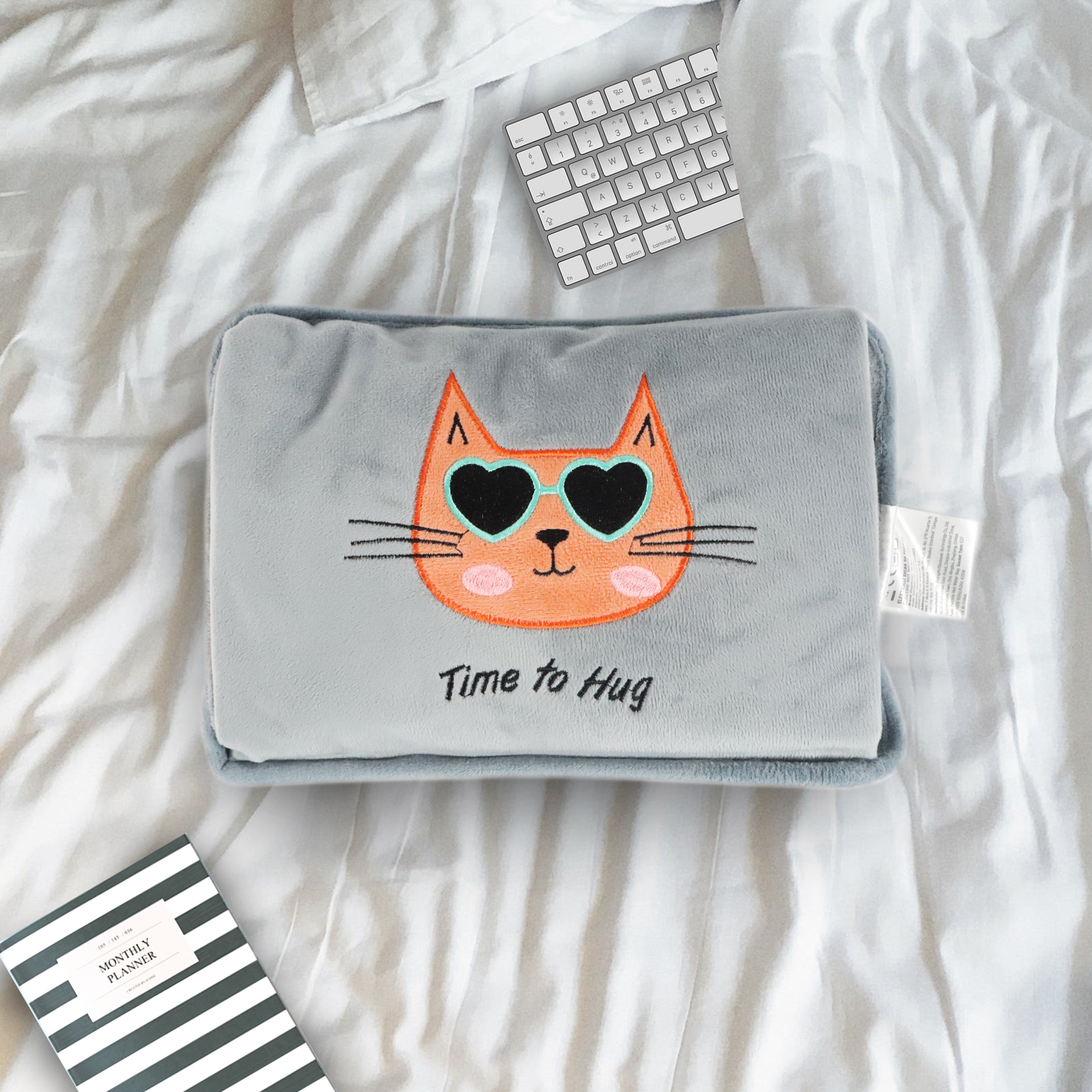 Almohada gris con cara de gato, gafas de sol de corazón, sobre cama con teclado y agenda.