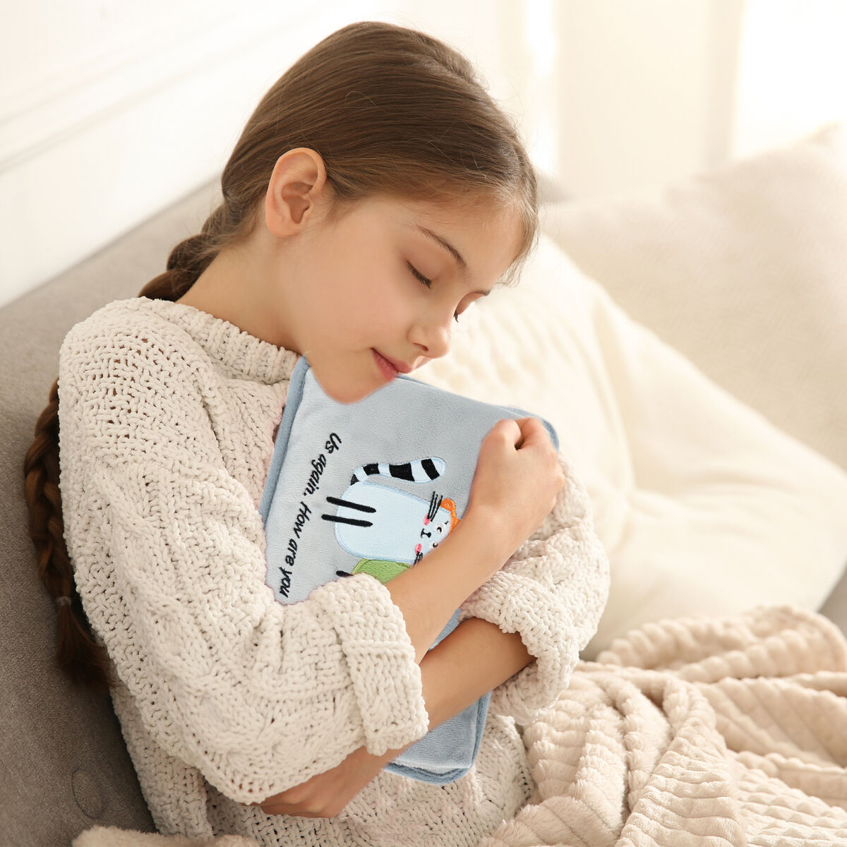Una niña con trenza abraza una bolsa de agua caliente azul con diseño de gato.