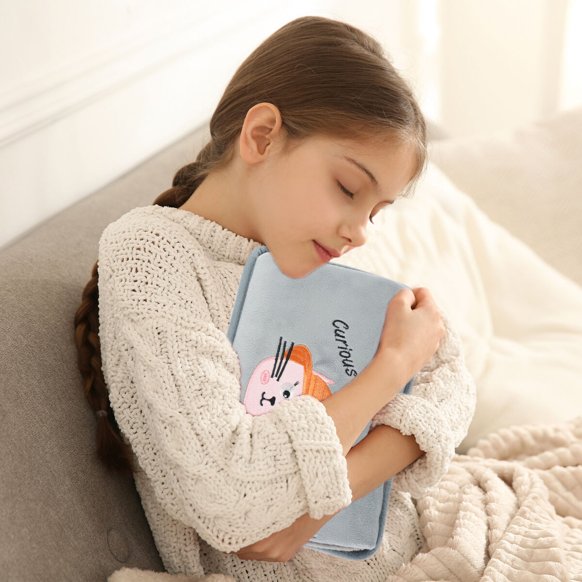 Una niña con trenza abraza una bolsa de agua caliente azul claro con diseño de conejito.