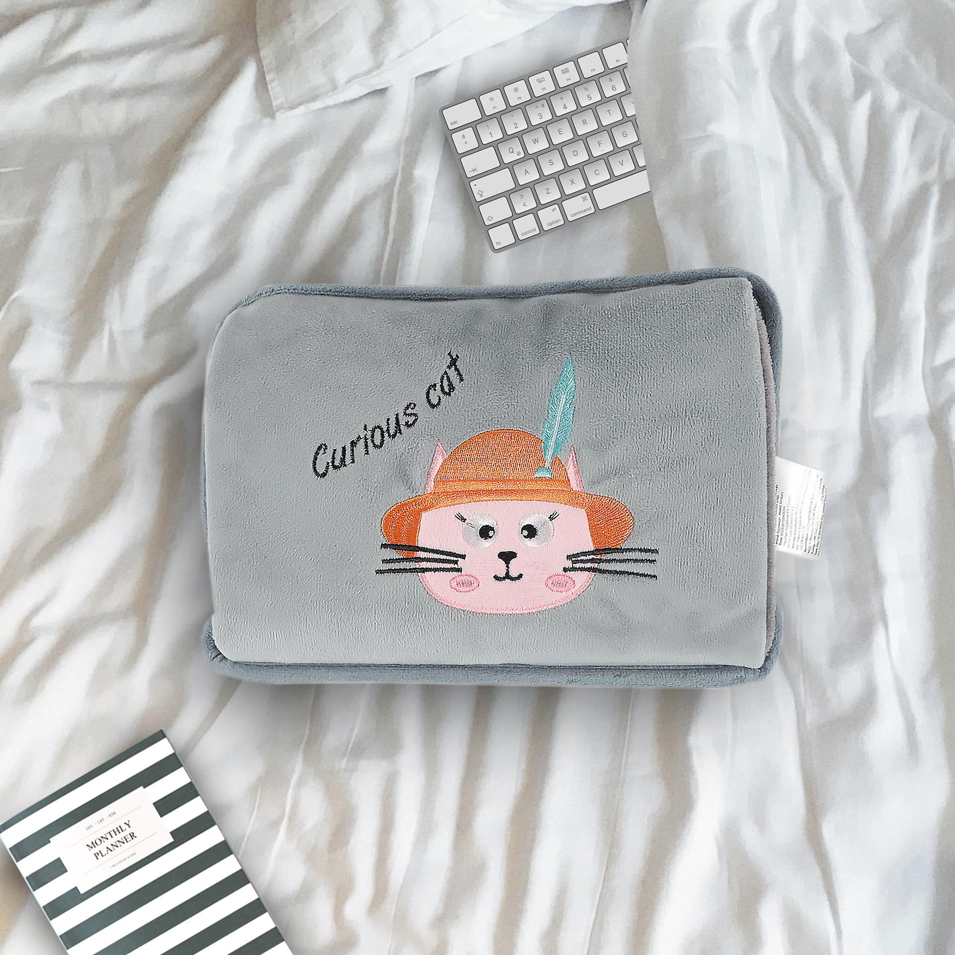Una almohada gris con un gato rosa con sombrero naranja y pluma yace sobre una cama blanca con teclado y planificador.