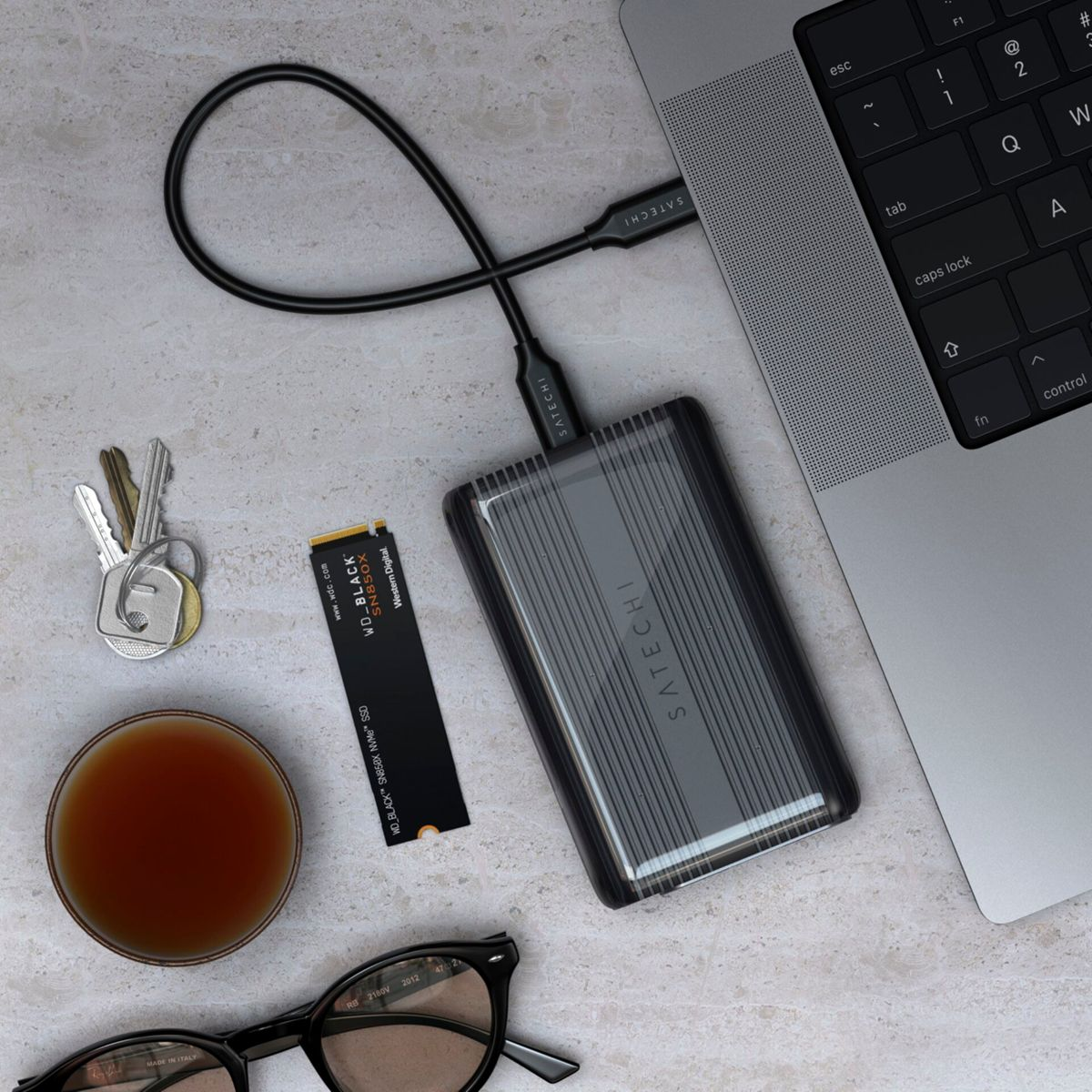 Disco esterno Satechi collegato al laptop, con SSD, chiavi, tazza e occhiali.