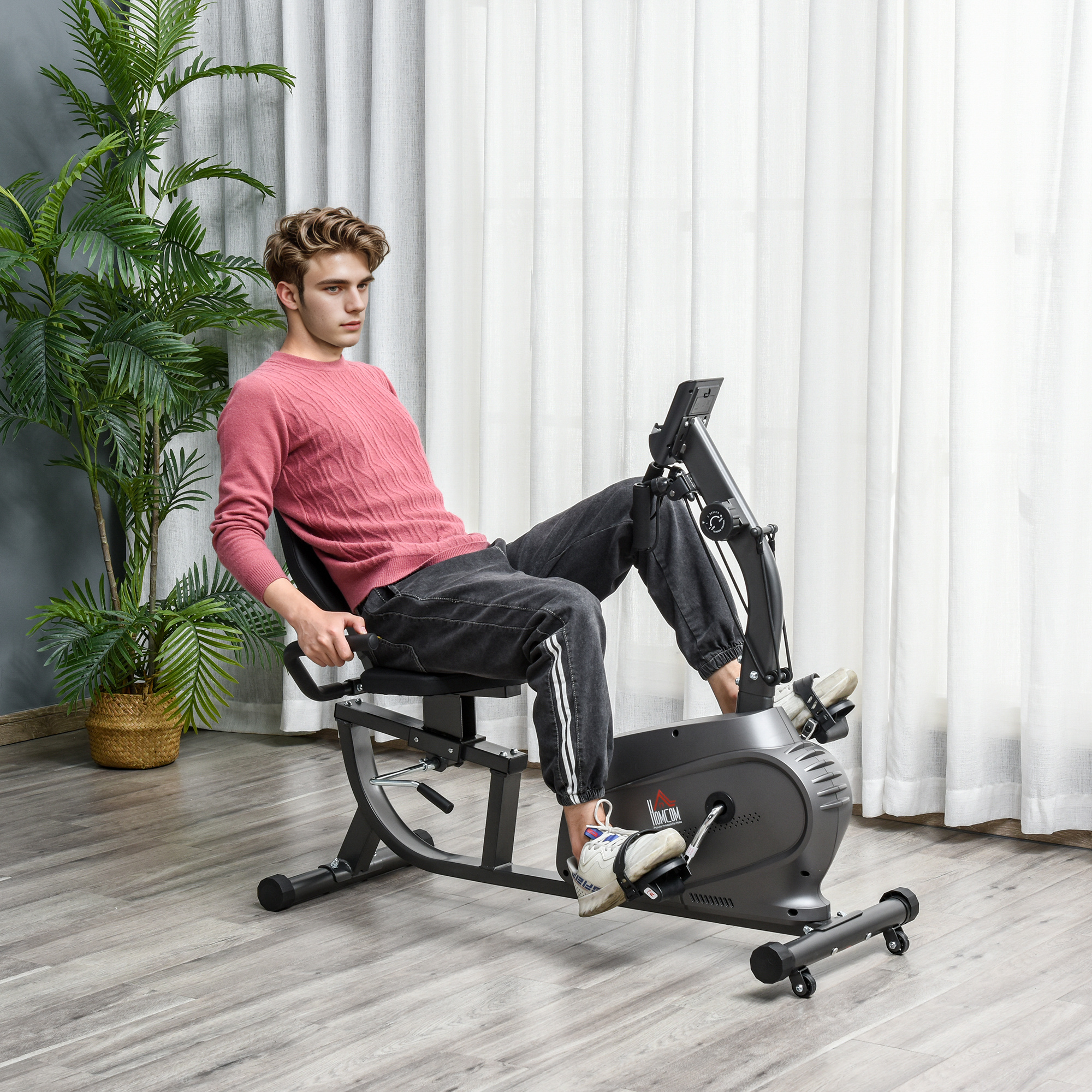 Un uomo in maglione rosa si allena su una cyclette reclinata davanti a una finestra con tende.