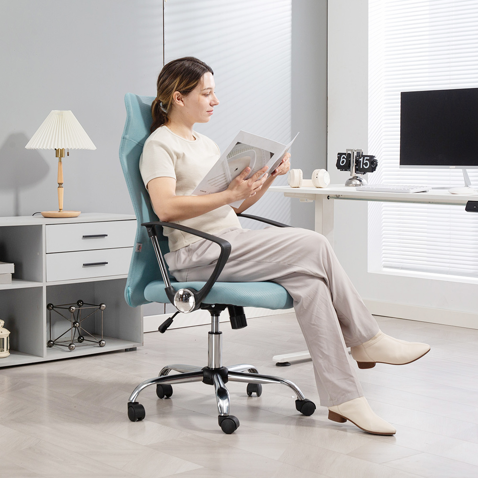 Una donna seduta su una sedia da ufficio legge una rivista. Una scrivania con monitor sullo sfondo.